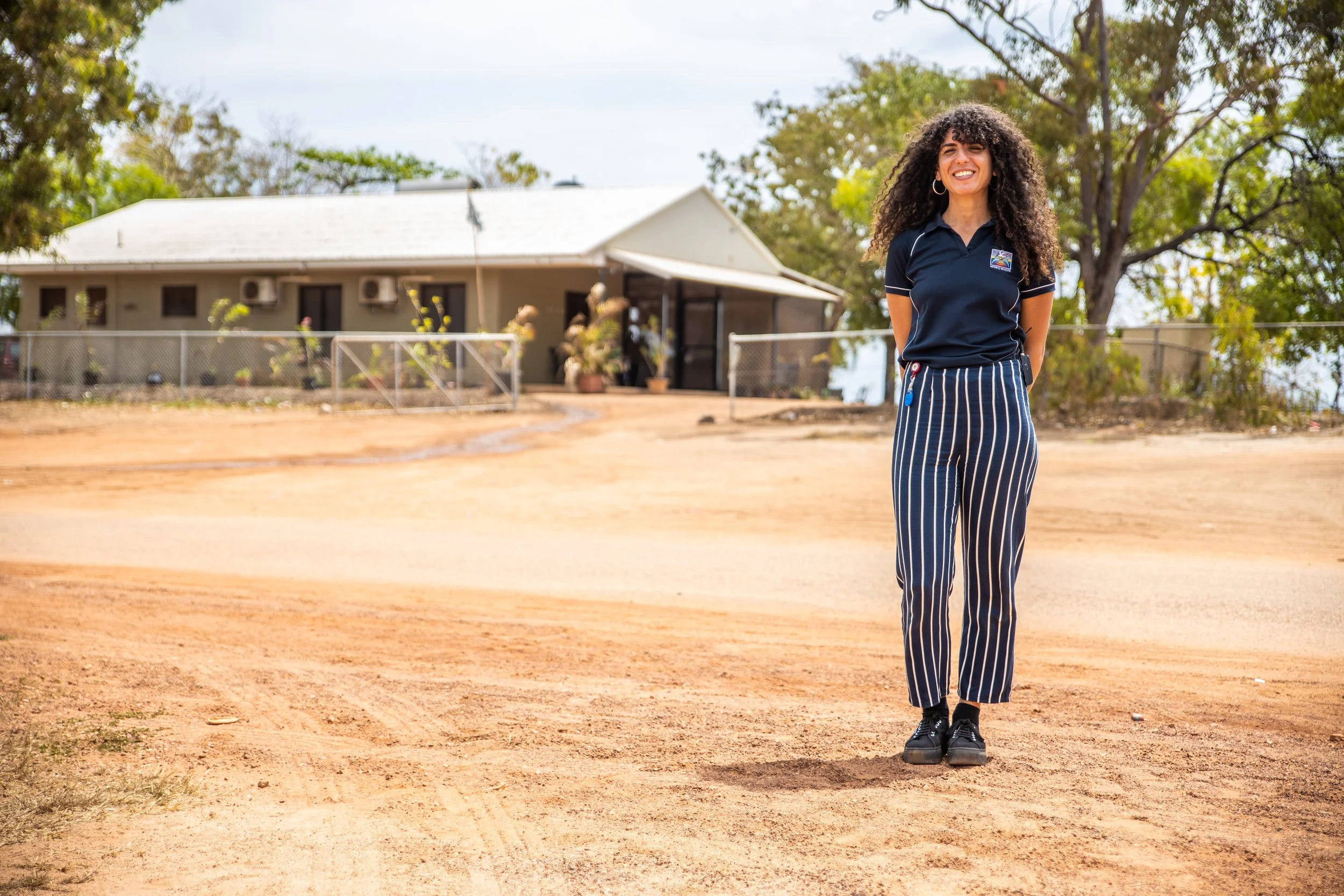 A woman with long curly hair smiling, standing on a dirt field with a house and trees in the background. She is wearing a dark blue polo shirt and striped pants.