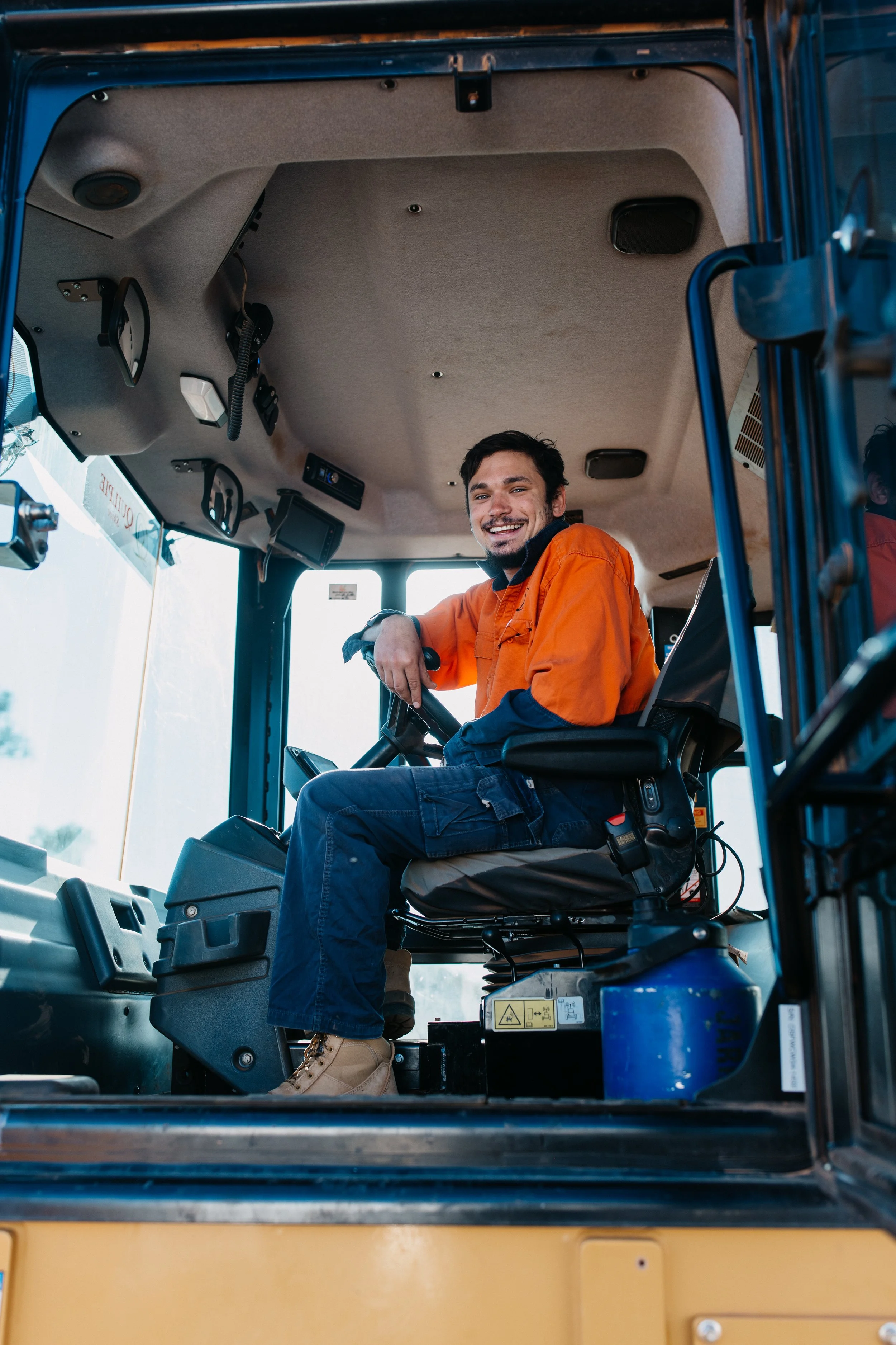 A smiling man in an orange work jacket and jeans sitting in the driver's seat of a construction vehicle, possibly a backhoe or excavator, inside the cabin with controls and equipment visible around him.