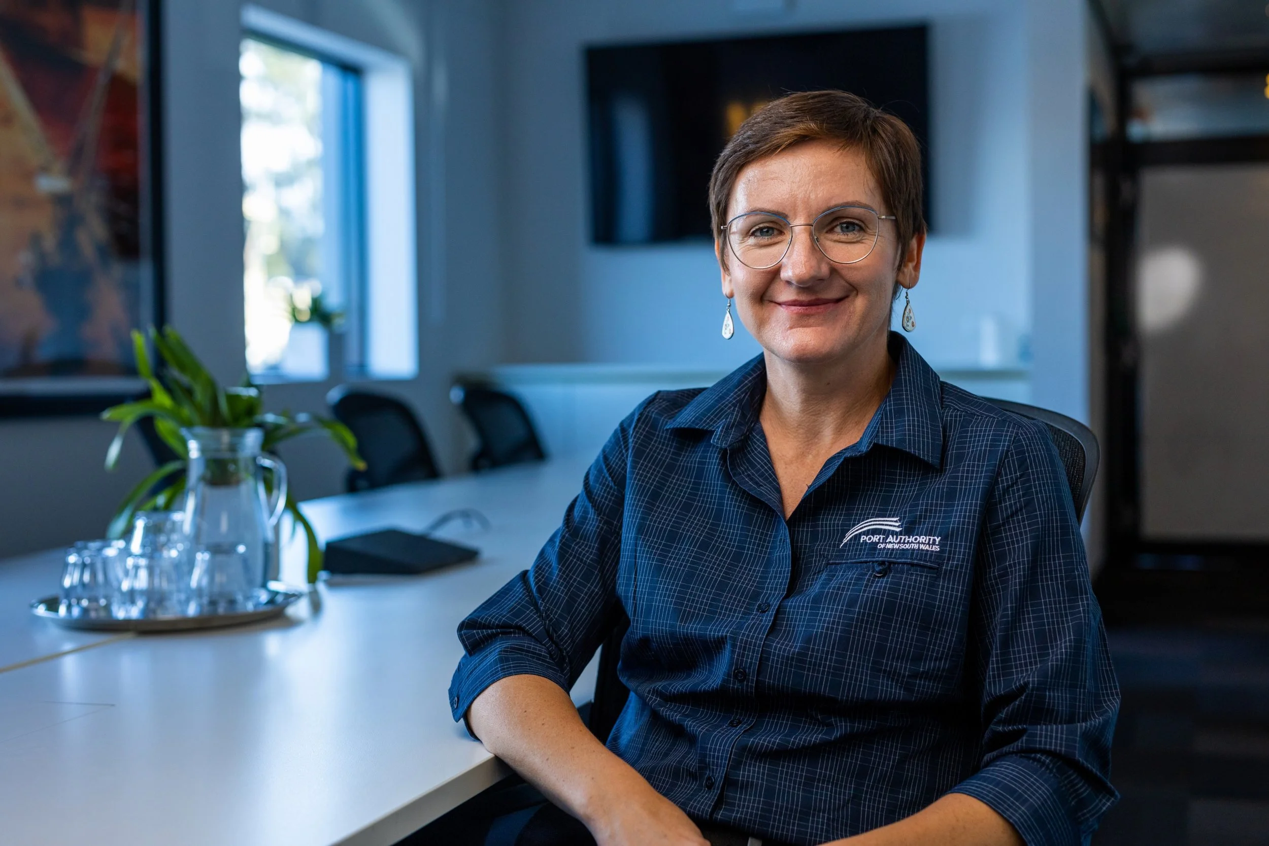 A woman with short brown hair, glasses, and earrings, wearing a navy blue shirt with a port authority logo, sitting at a conference table in a modern office.