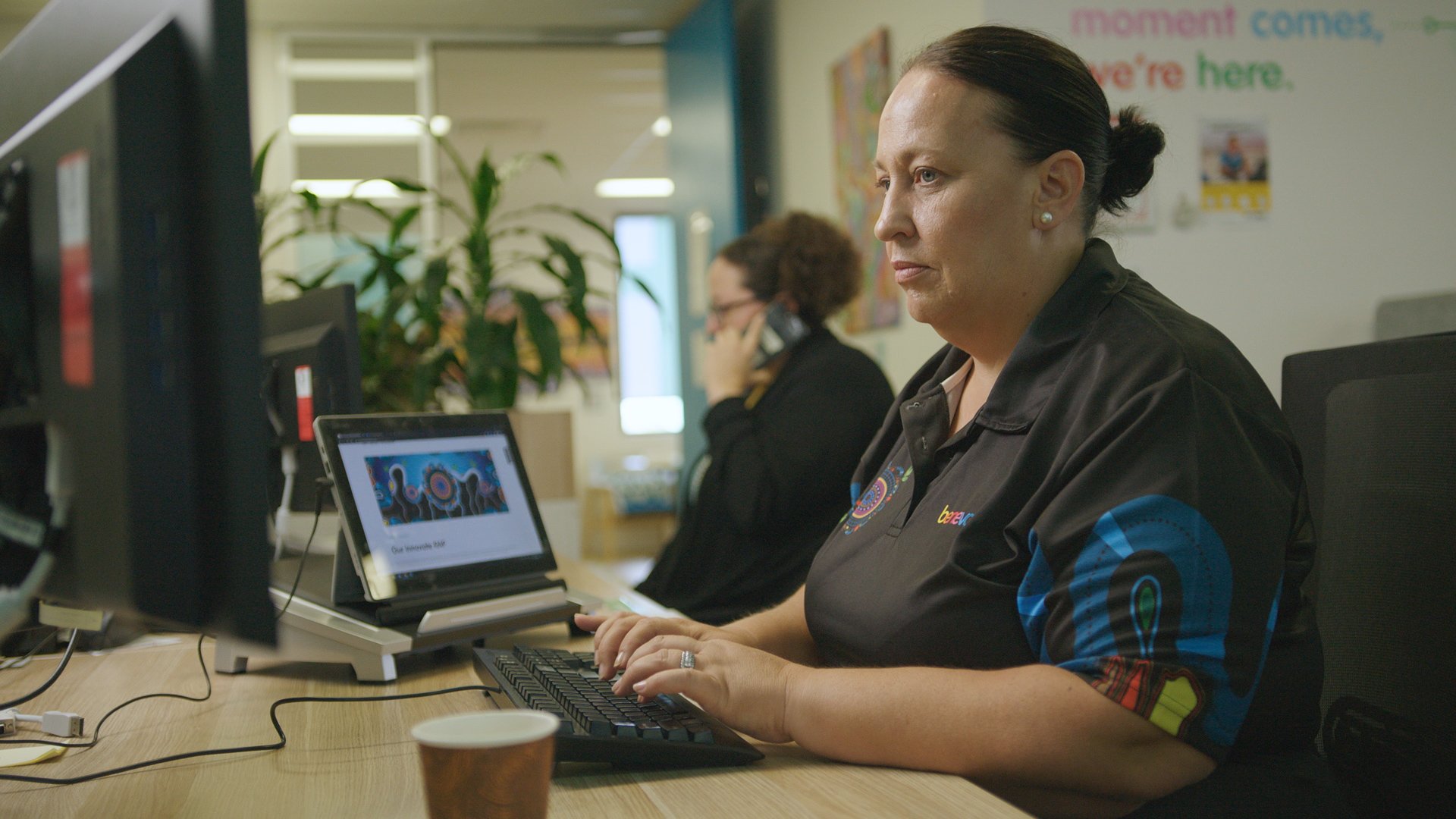 A woman working at a desk on a computer in an office with another woman on the phone in the background.