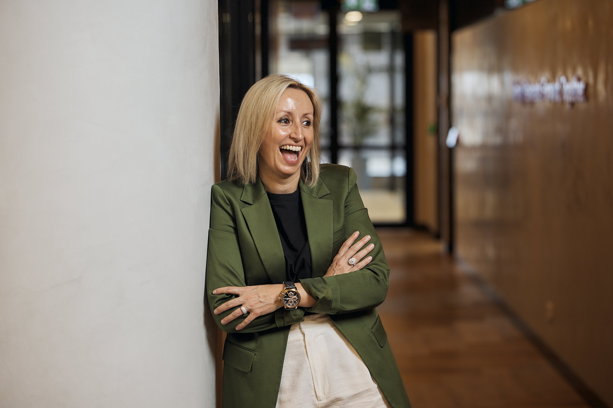 A woman with blonde hair wearing a green blazer and beige skirt, smiling and leaning against a wall in a modern interior space.