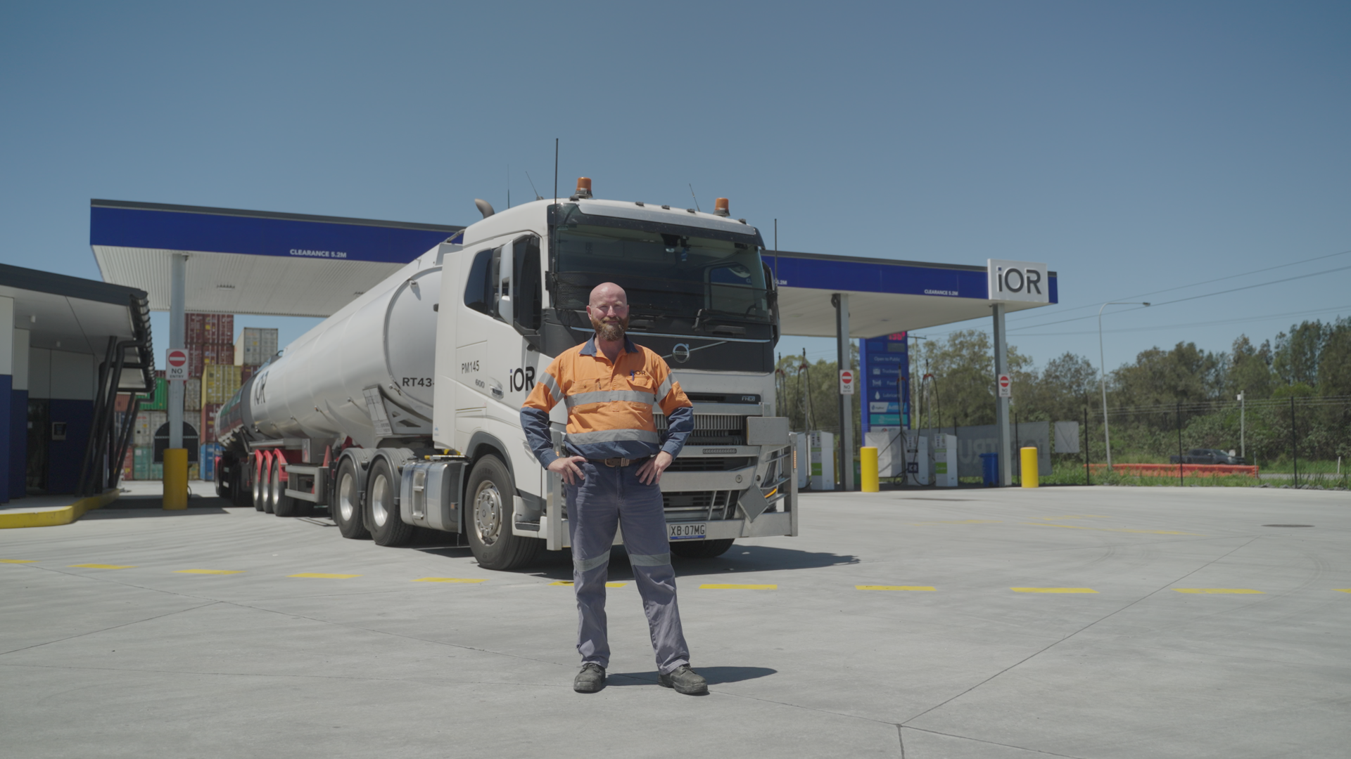 A worker in safety gear stands in front of a tanker truck at a fuel station on a clear, sunny day.
