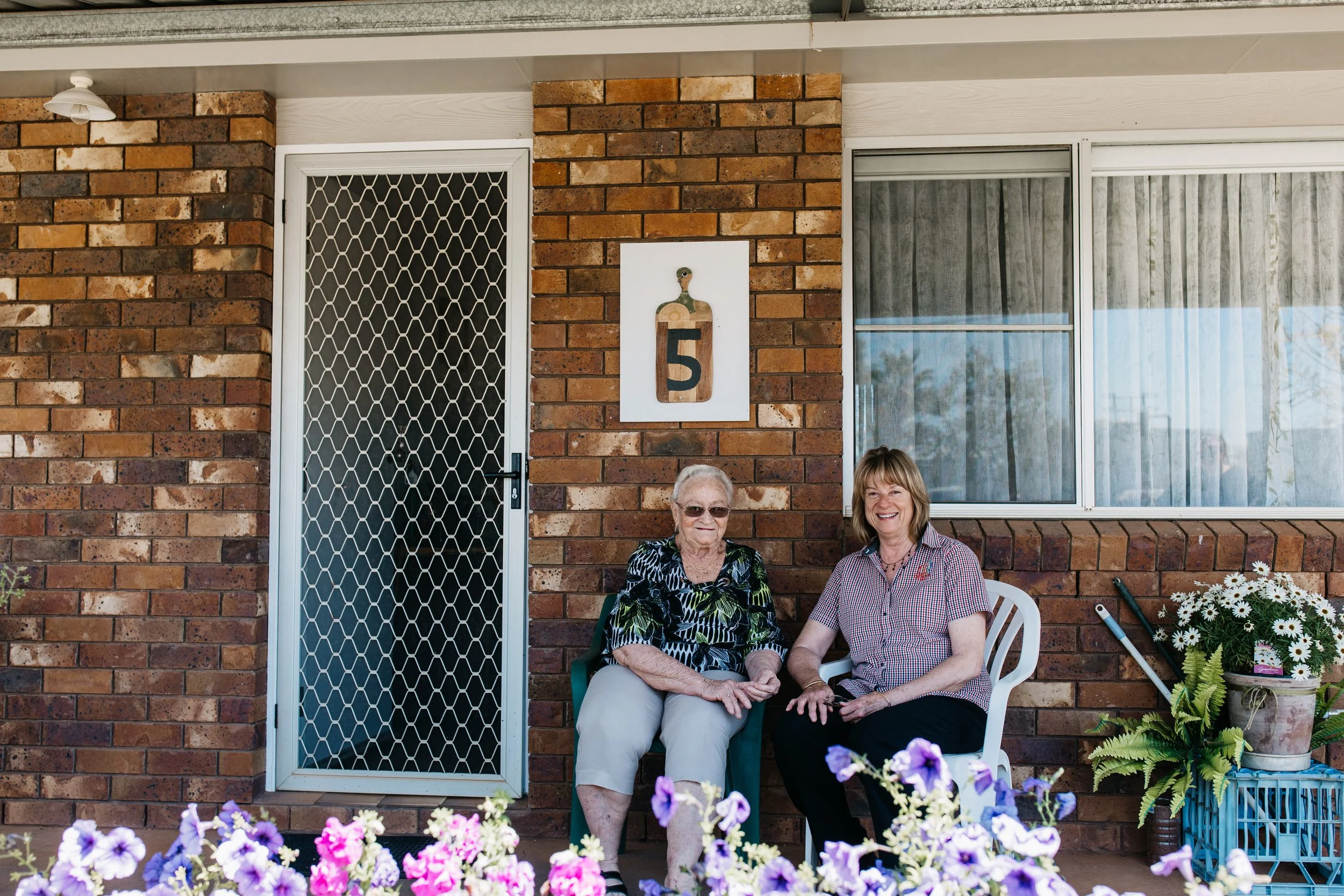 Two women sitting on patio chairs in front of a brick house, surrounded by colorful flowers.