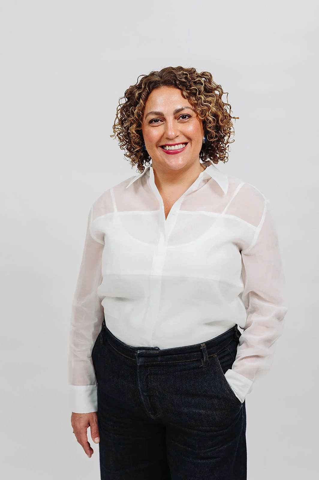 A woman with curly hair wearing a white shirt and jeans, smiling, leaning on a white picket fence outdoors.