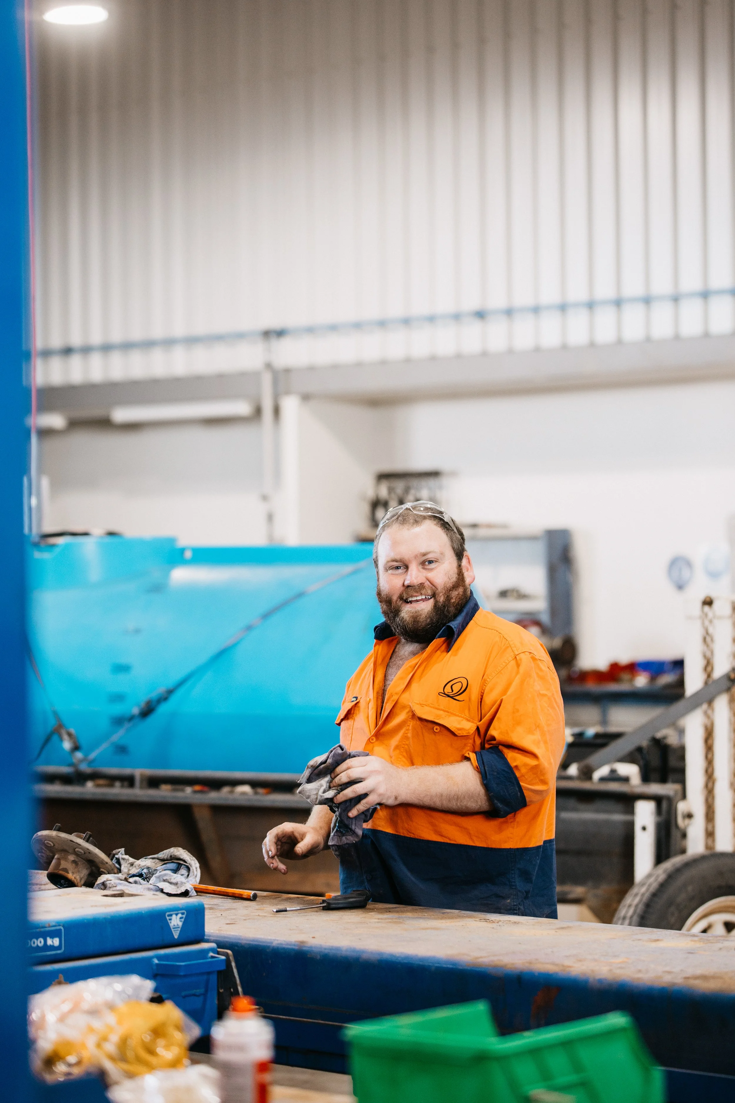 A smiling man with a beard, wearing an orange and navy work uniform, standing in an industrial workshop. He is holding a cloth and working at a cluttered workbench with tools and materials. In the background, there is a large blue object, possibly a boat, and the workshop has metal walls and equipment.