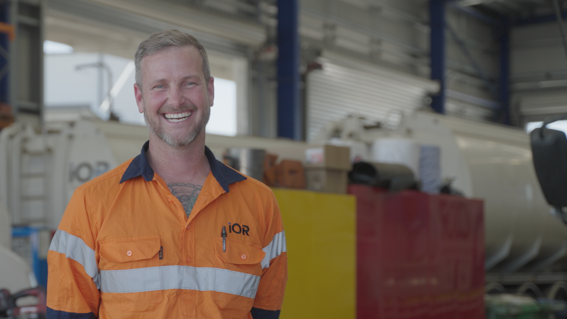 A smiling man in an orange safety uniform with the logo 'IOR' stands in an industrial or warehouse setting, with machinery and storage boxes in the background.