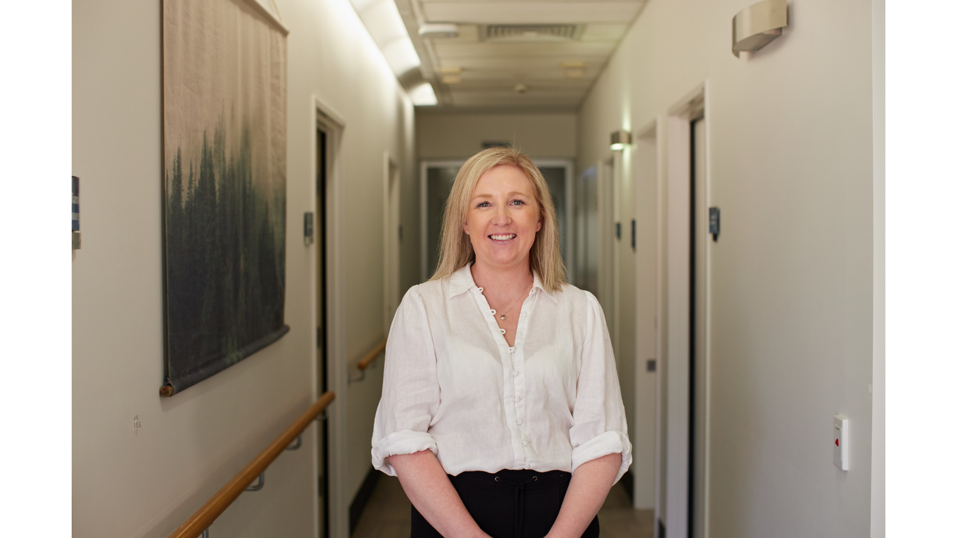 A woman with blonde hair smiling in a hallway, wearing a white blouse and black pants.