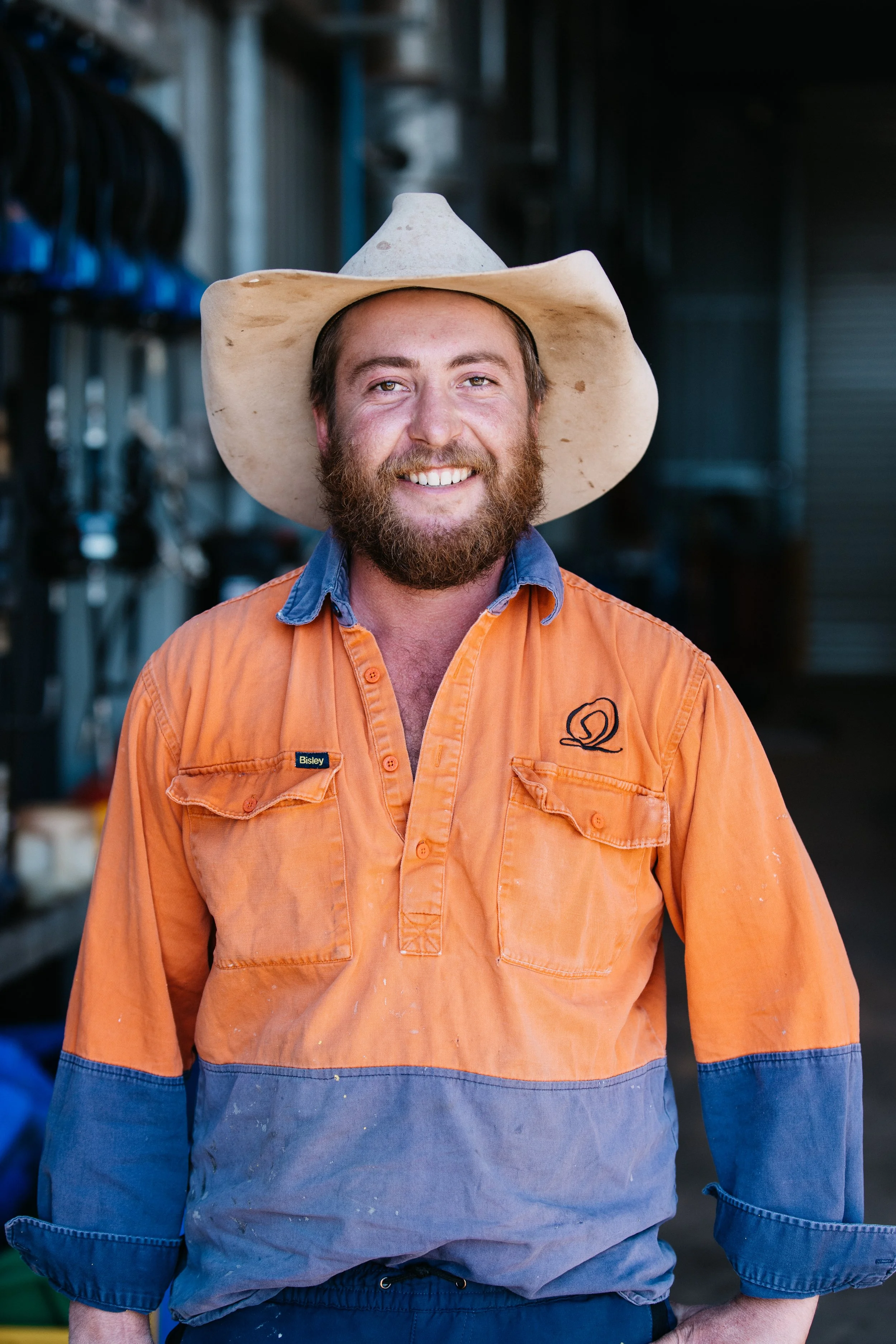 Portrait of a man smiling, wearing a wide-brimmed hat, orange and blue work shirt with a logo, standing in a workshop or garage.