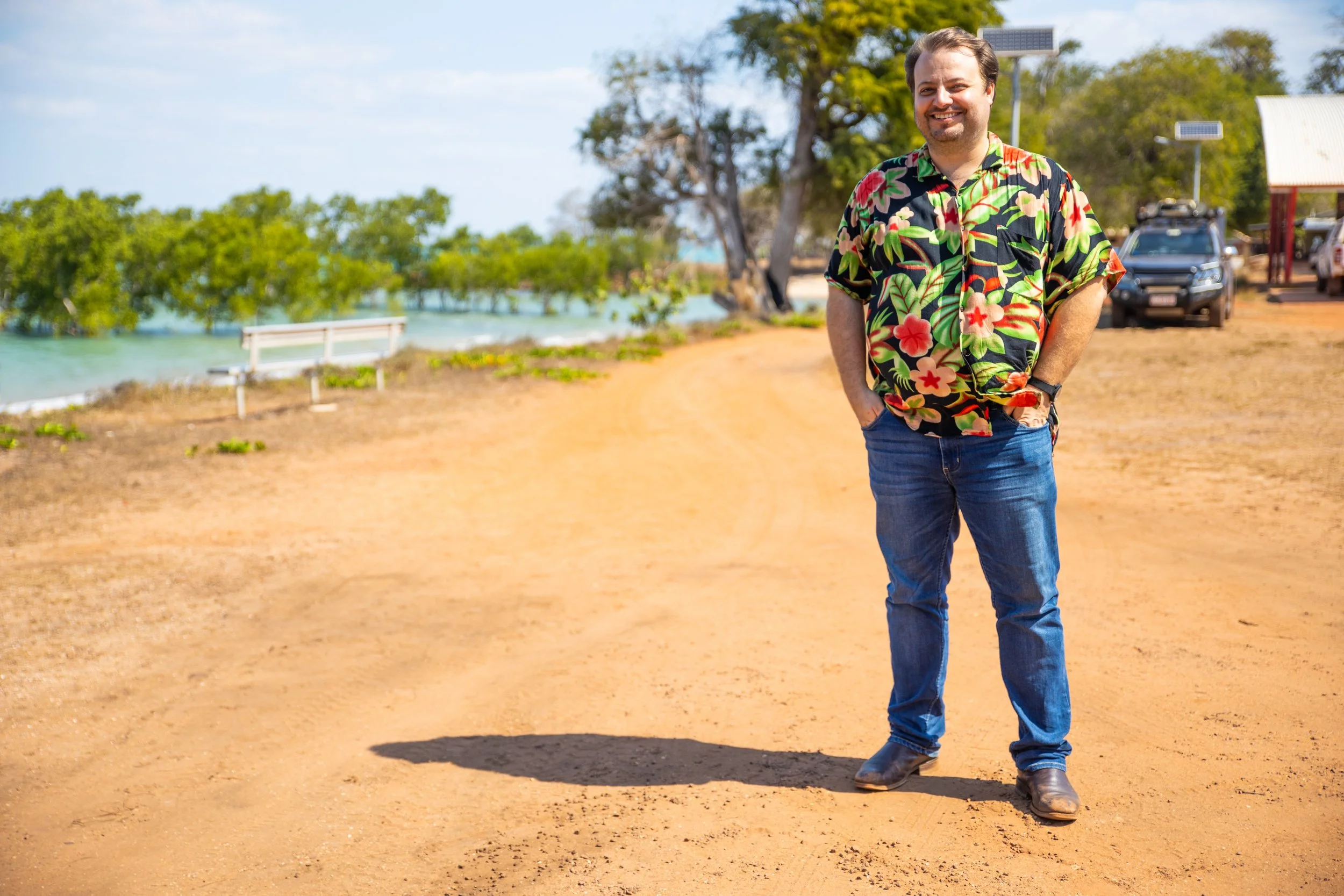 A man standing on a dirt path near water, wearing a colorful Hawaiian shirt and jeans, smiling, with trees, a bench, and parked vehicles in the background.