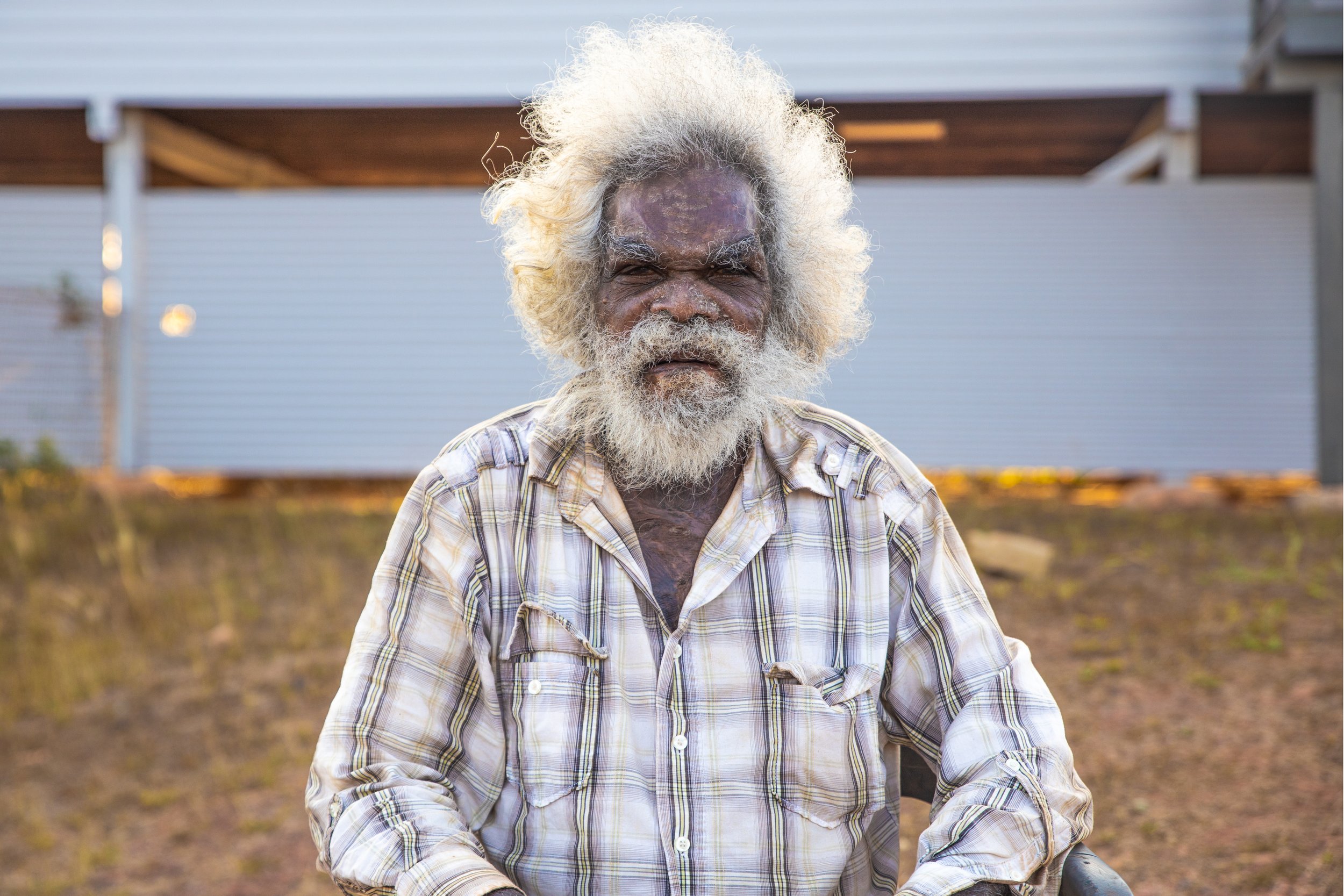 An elderly man with white, curly hair and beard, wearing a plaid shirt, sits outdoors with a blurred background of a modern building and garden.