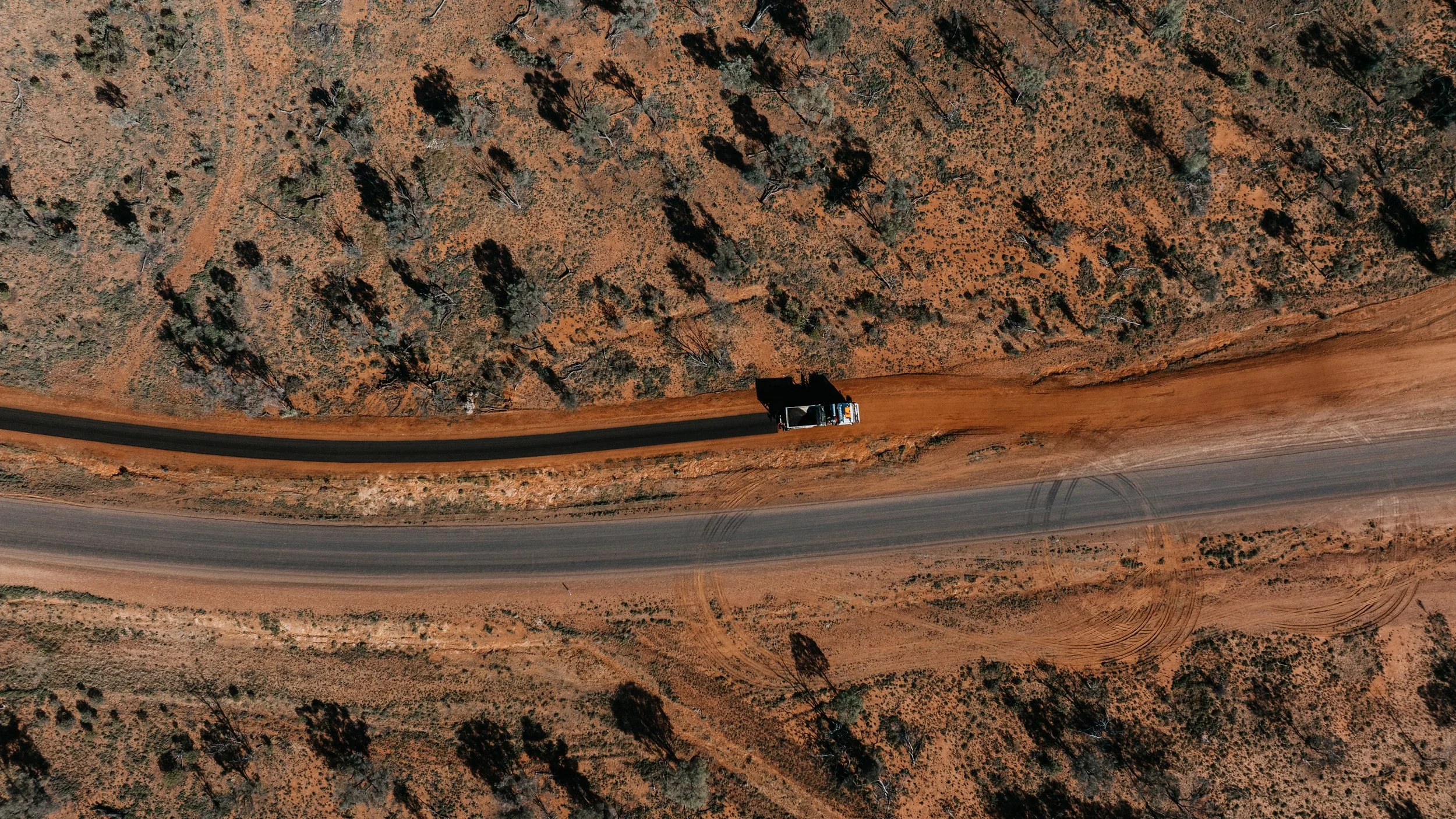 An aerial view of a dirt road cutting through a semi-arid landscape with sparse trees and bushes, and a pickup truck traveling along the dirt road.
