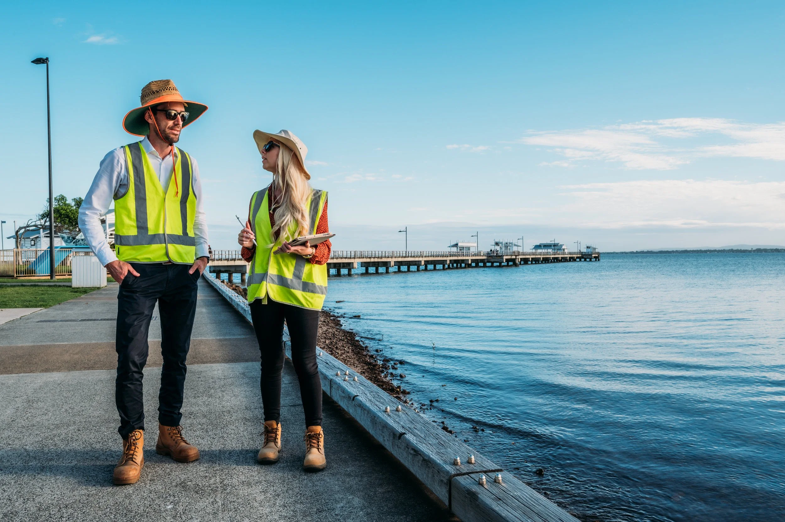 Two workers in yellow safety vests and boots standing by a waterfront pier, discussing with one holding a clipboard and pen, under a clear blue sky.