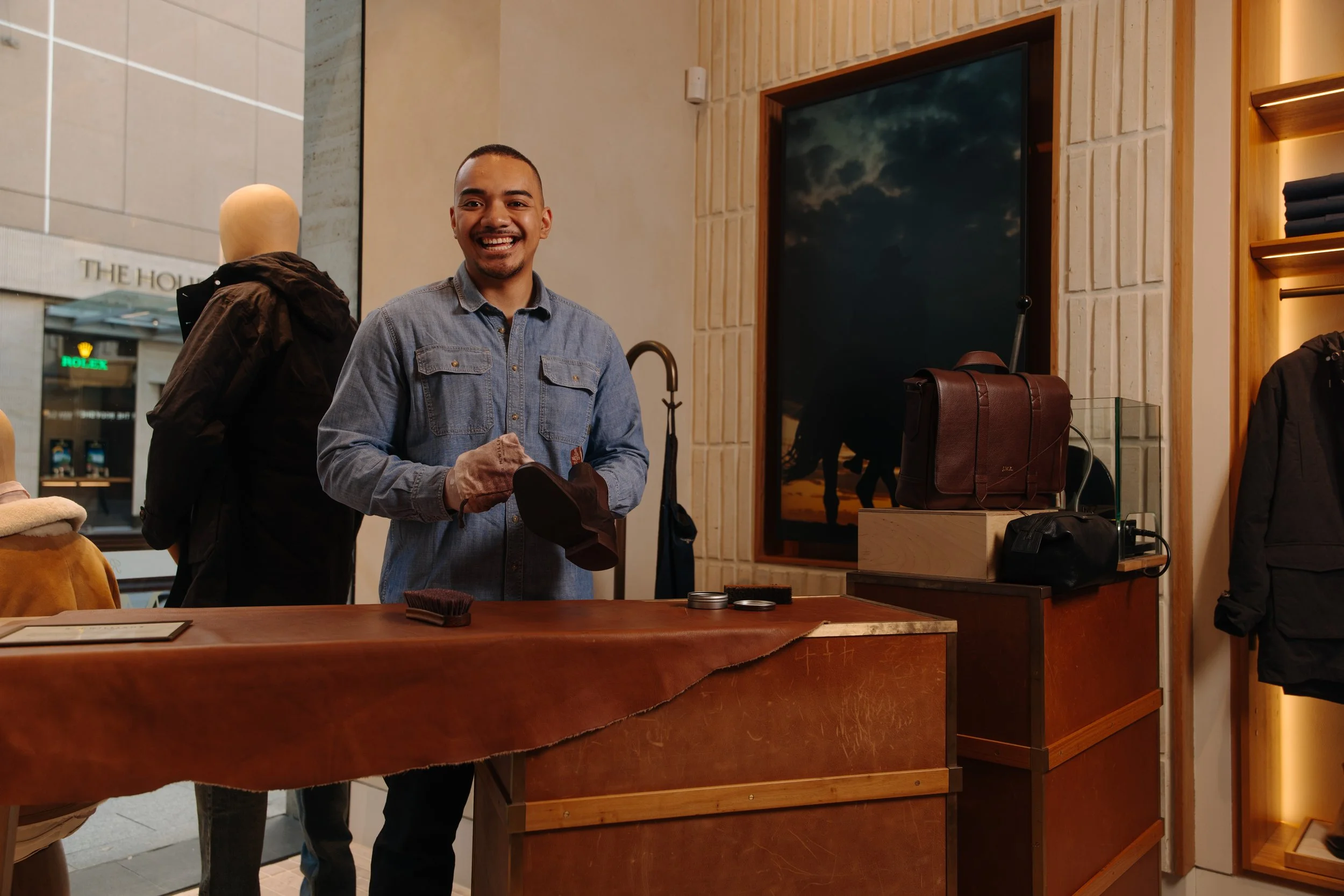 A smiling man in a denim shirt standing behind a wooden counter inside a retail store, with a large window and modern decor in the background.