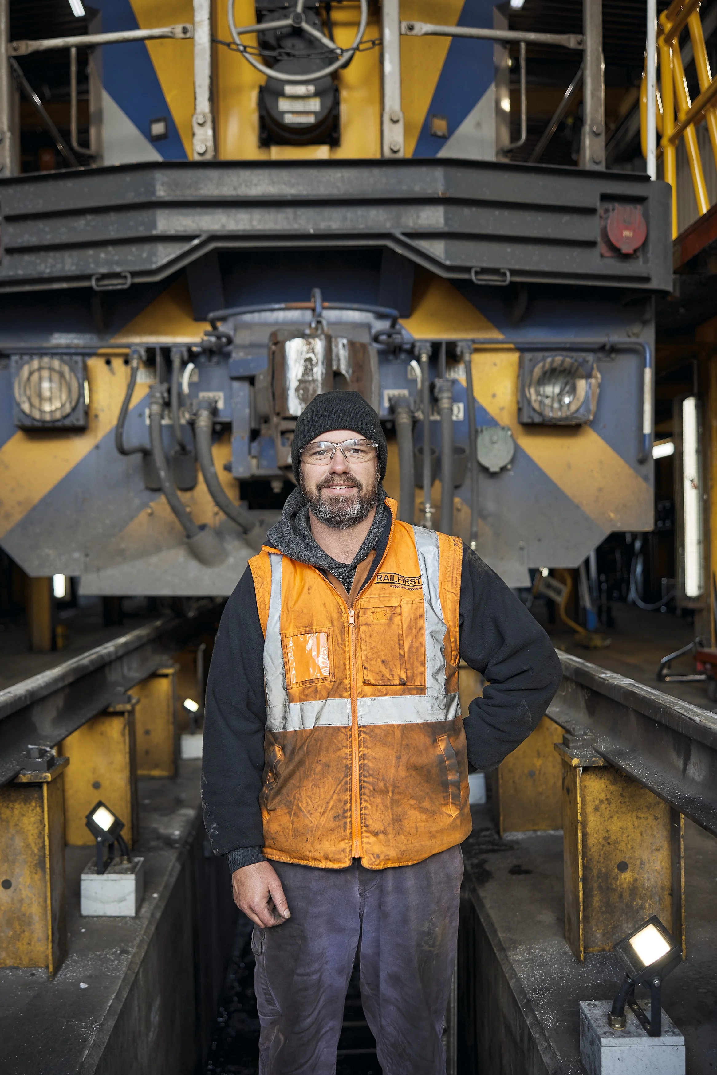 A man wearing safety glasses, a black beanie, a gray hoodie, and an orange safety vest stands in front of a large industrial machine with yellow and black hazard stripes, various pipes, and equipment in an industrial setting.