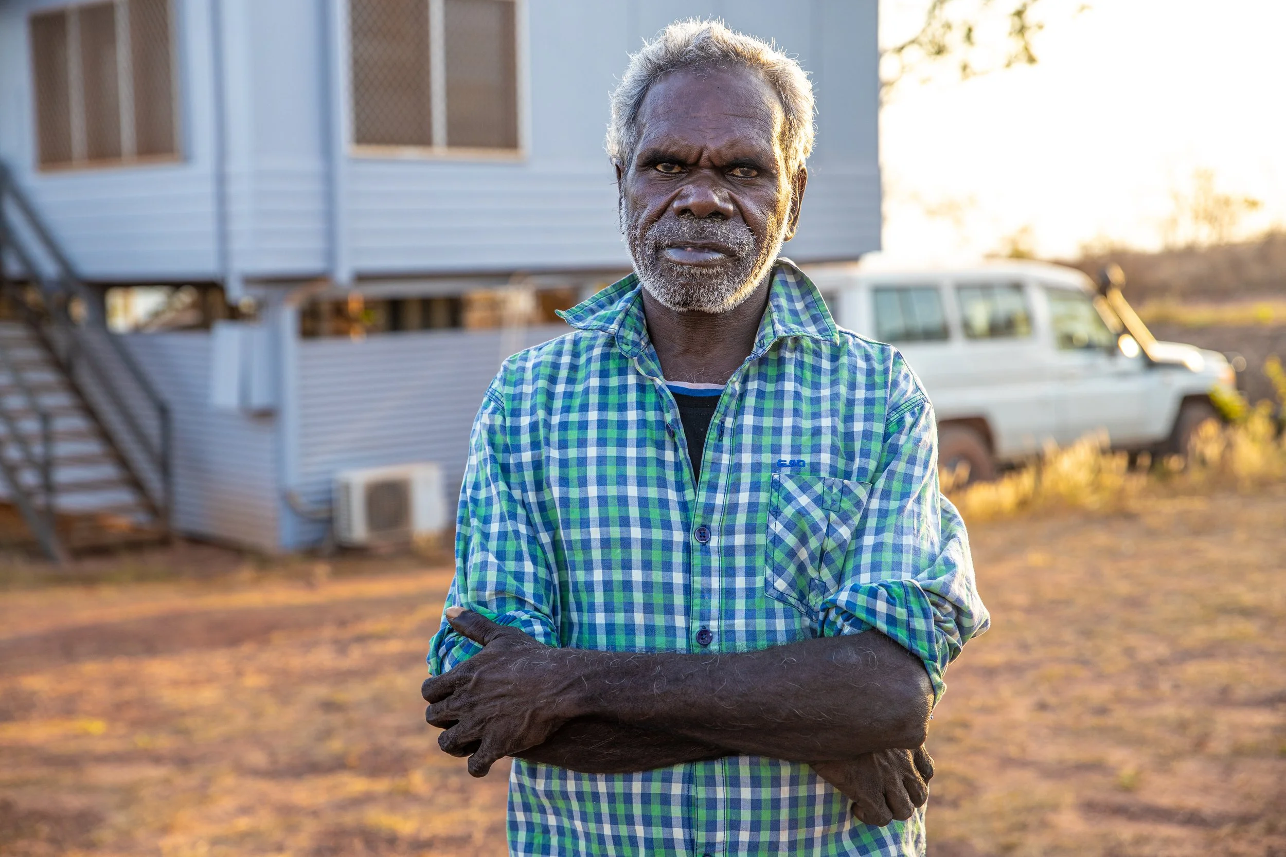 An elderly man with gray hair and a beard, wearing a green and blue checkered shirt, stands outdoors with his arms crossed, in front of a mobile home and a vehicle, during sunset.