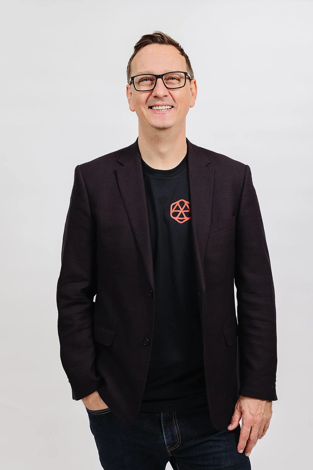 A smiling man in a black blazer and T-shirt standing outdoors with greenery and a blurred background.