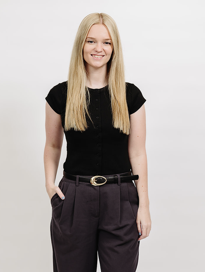 A young woman with long blonde hair, wearing a black top and gray pants, posing outdoors next to a white picket fence, smiling, with trees and a building in the background.