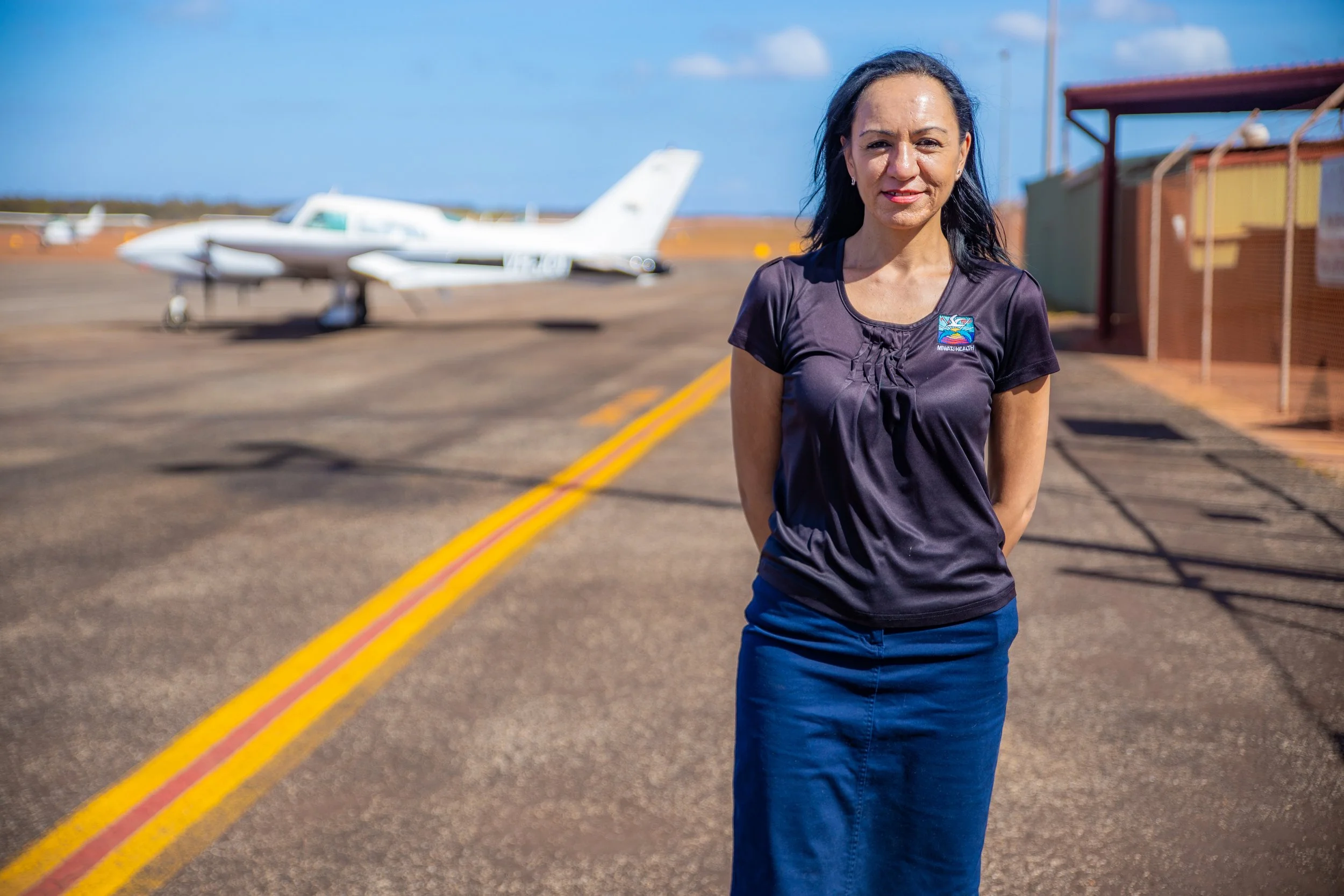 A woman stands outside near an airplane on an airfield, with a blue sky and some clouds in the background.
