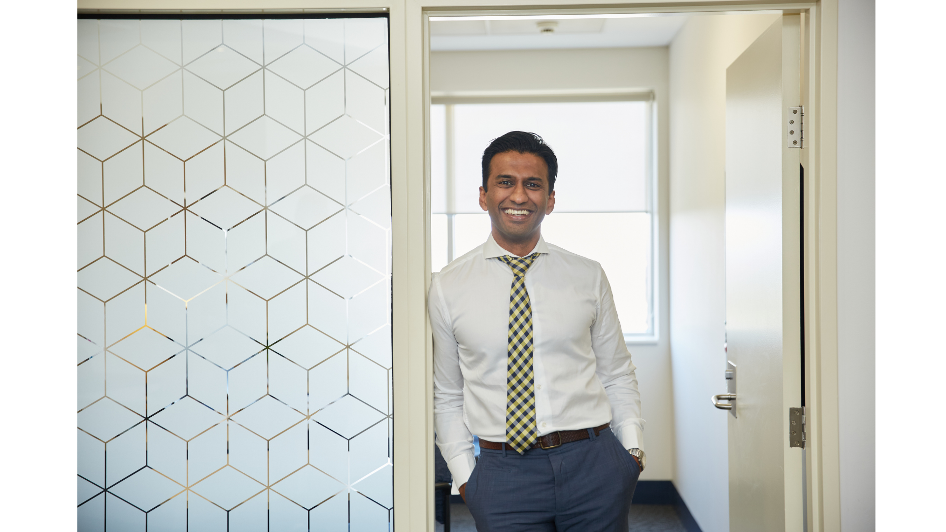 A man in business attire standing in an office doorway, smiling, wearing a white shirt, plaid tie, and blue pants with hands in pockets, with a patterned partition on the left and windows in the background.