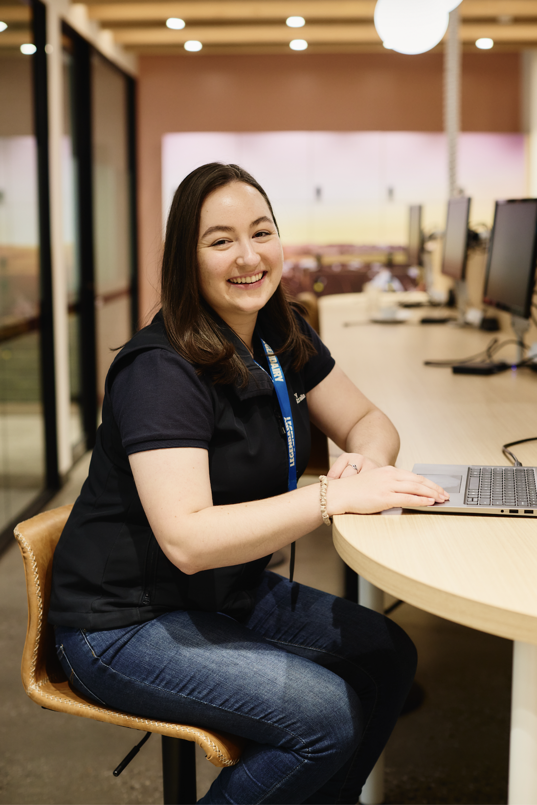 A young woman with dark hair and light skin sitting at a round table with a laptop in a modern, well-lit office space, smiling at the camera.