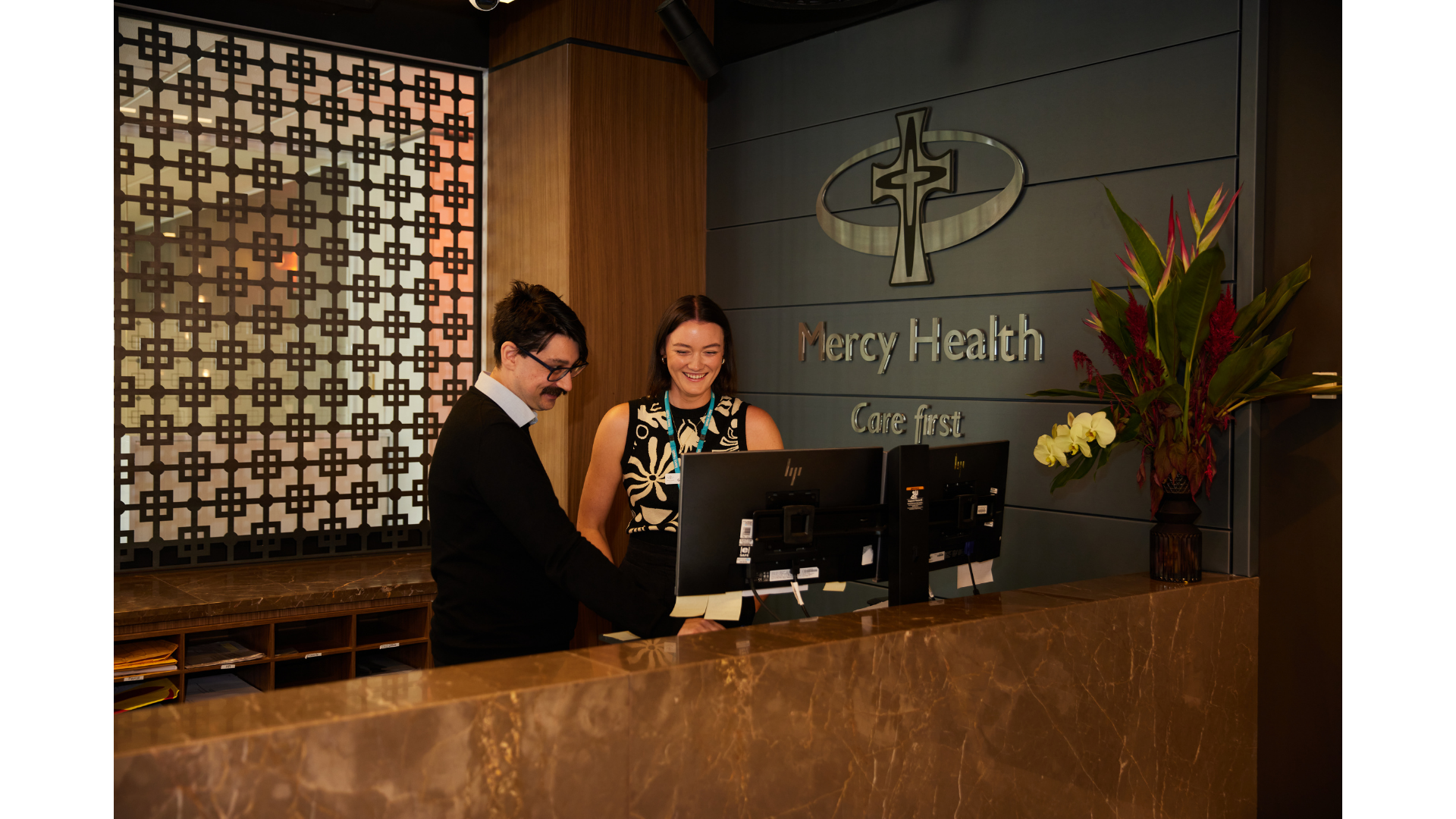 Reception area at Mercy Health with two staff members reviewing information at the front desk. There are two computer monitors, a decorative wall sign with a cross, and a large flower arrangement.