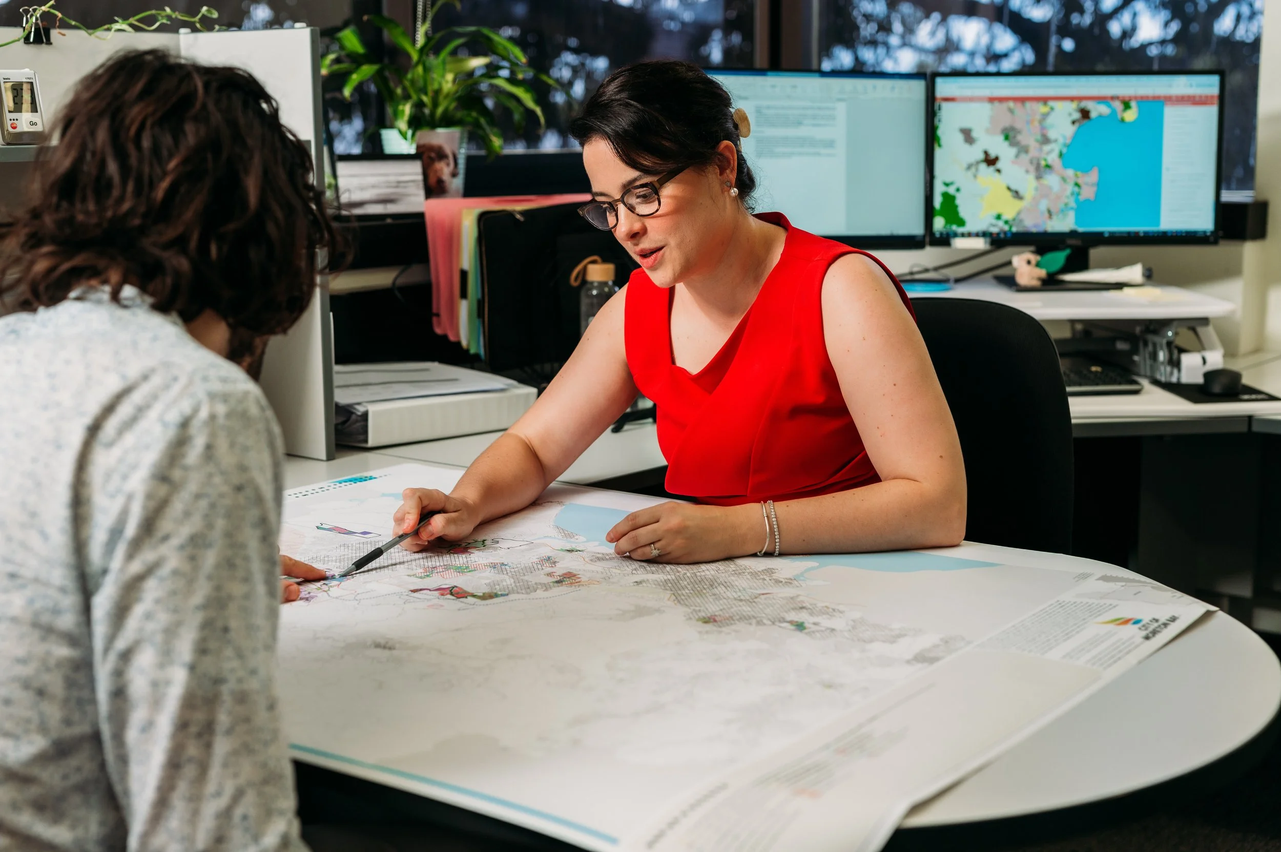 Two women are sitting at a desk reviewing a detailed map, with multiple computer monitors in the background displaying information and maps, in an office setting.