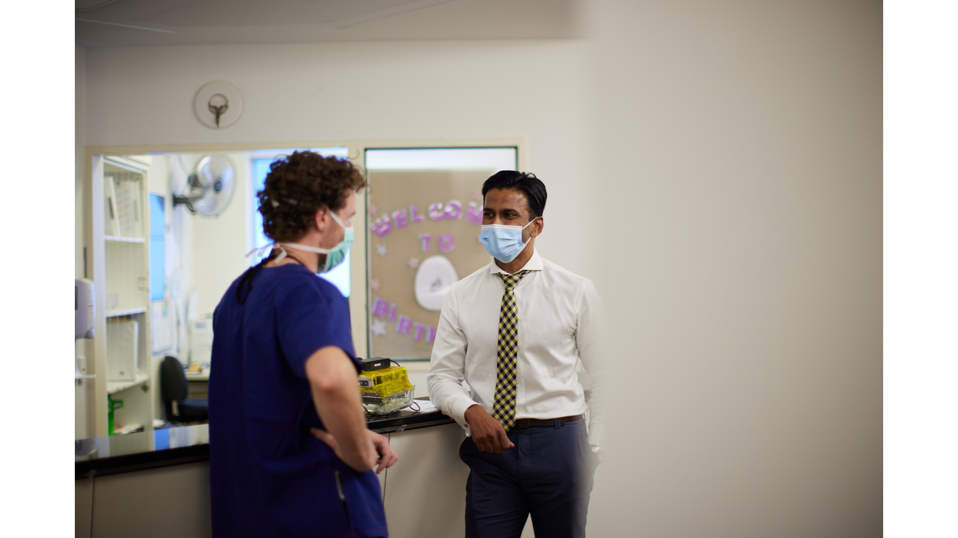 Two healthcare professionals, both wearing face masks, are having a conversation in a hospital or clinic. One is a woman in scrubs, and the other is a man dressed in formal attire with a patterned tie. The background includes medical equipment and a decorative wall sign.