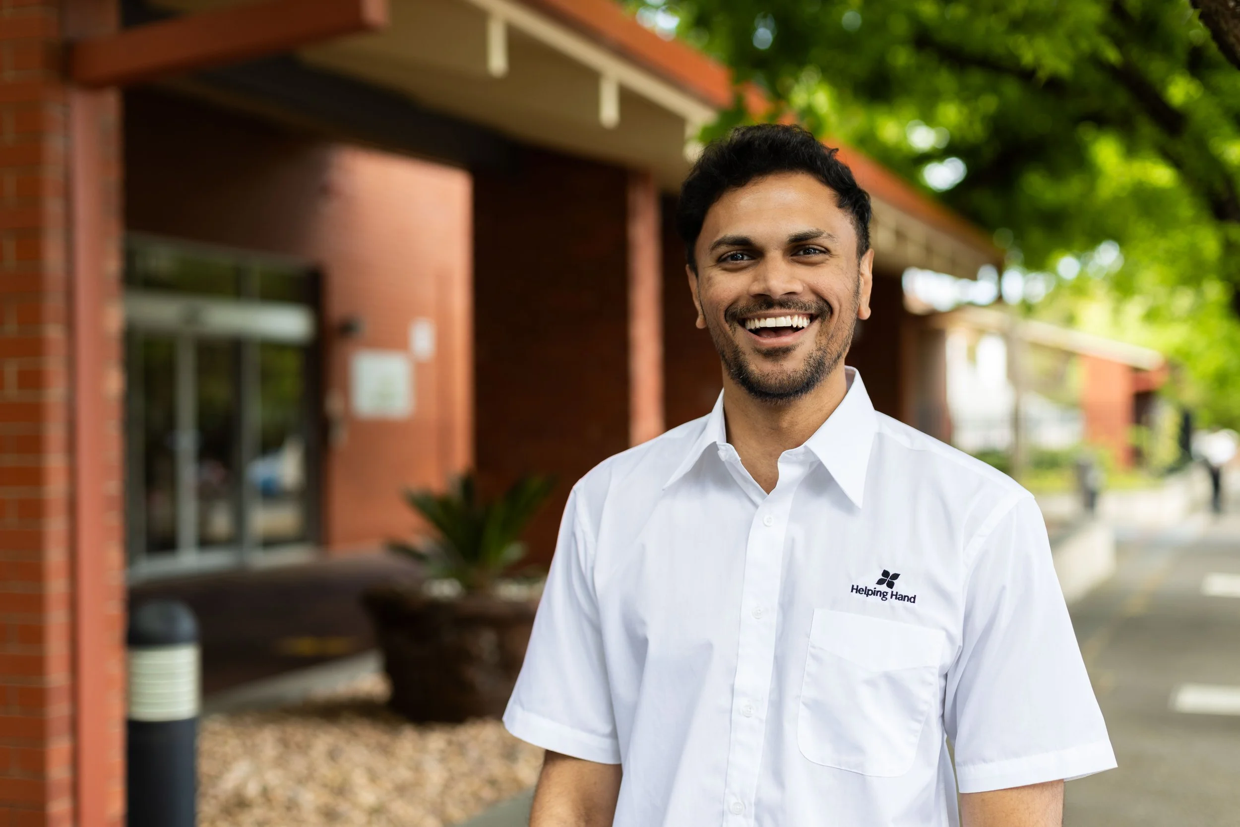 A smiling man in a white shirt with a 'Helping Hand' logo standing outside in front of a building with green trees overhead.