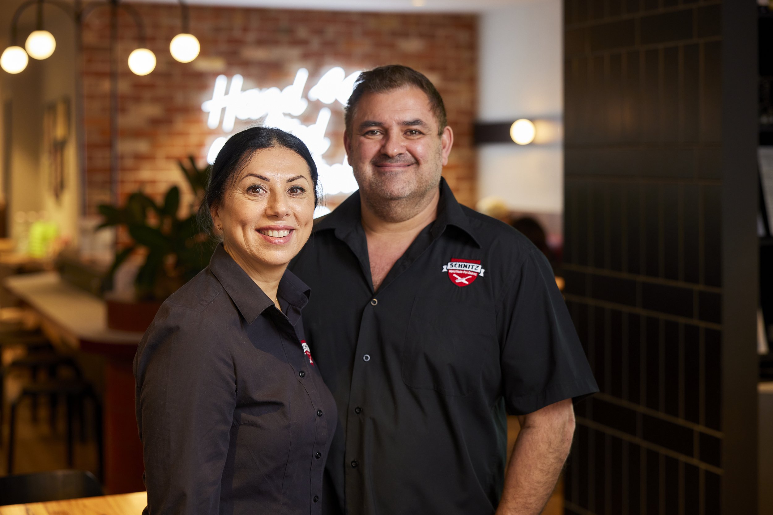 A man and a woman smiling at the camera in a restaurant or cafe with a brick wall and hanging lights in the background.