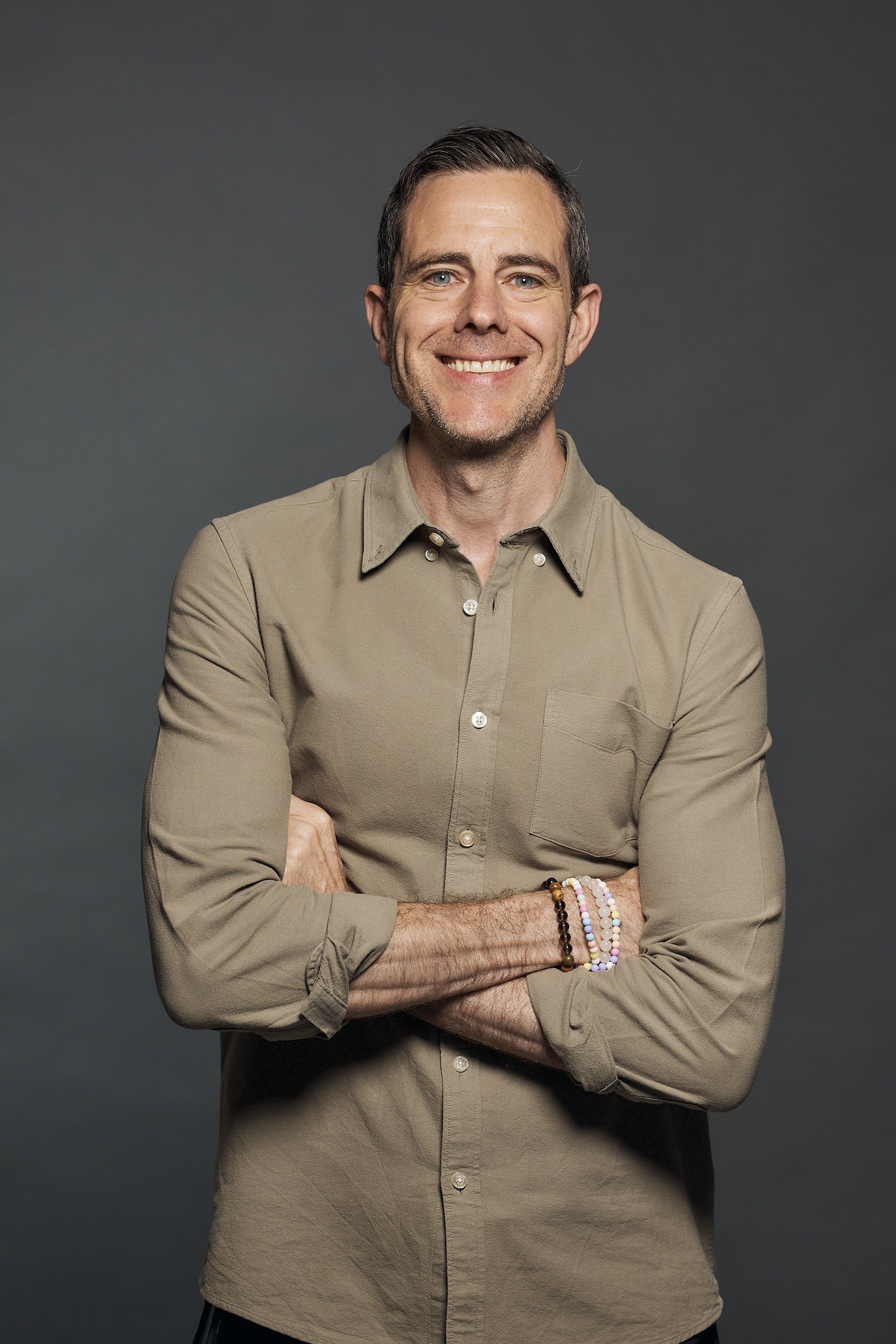 A smiling man with short dark hair and blue eyes, wearing a beige button-up shirt and multiple colorful beaded bracelets on his left wrist, standing with arms crossed against a dark gray background.