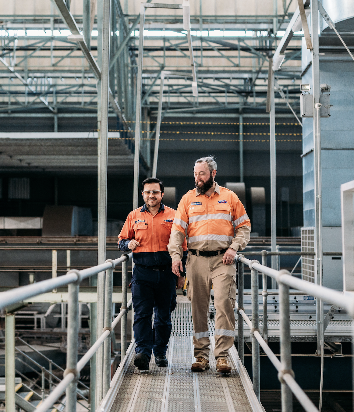 Two workers walking on a metal pathway inside an industrial facility, wearing safety uniforms and smiling.