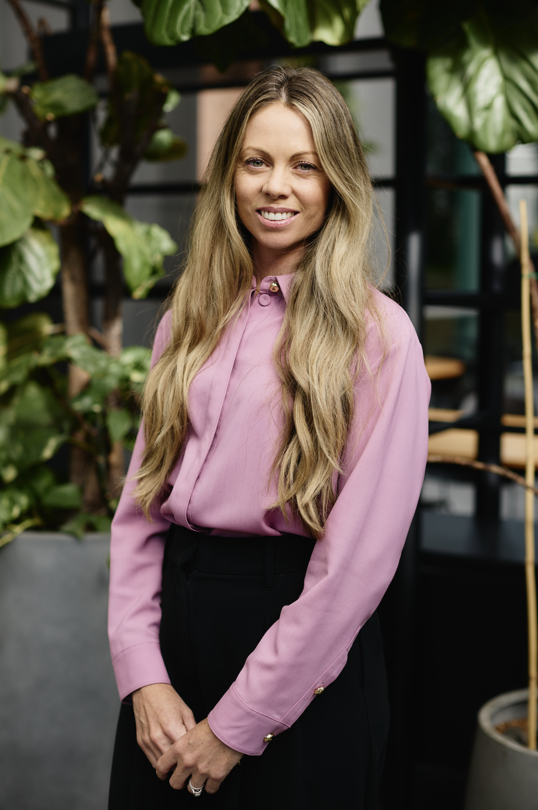 A woman with long blonde hair wearing a pink blouse and black pants, standing in front of green plants and modern shelves.