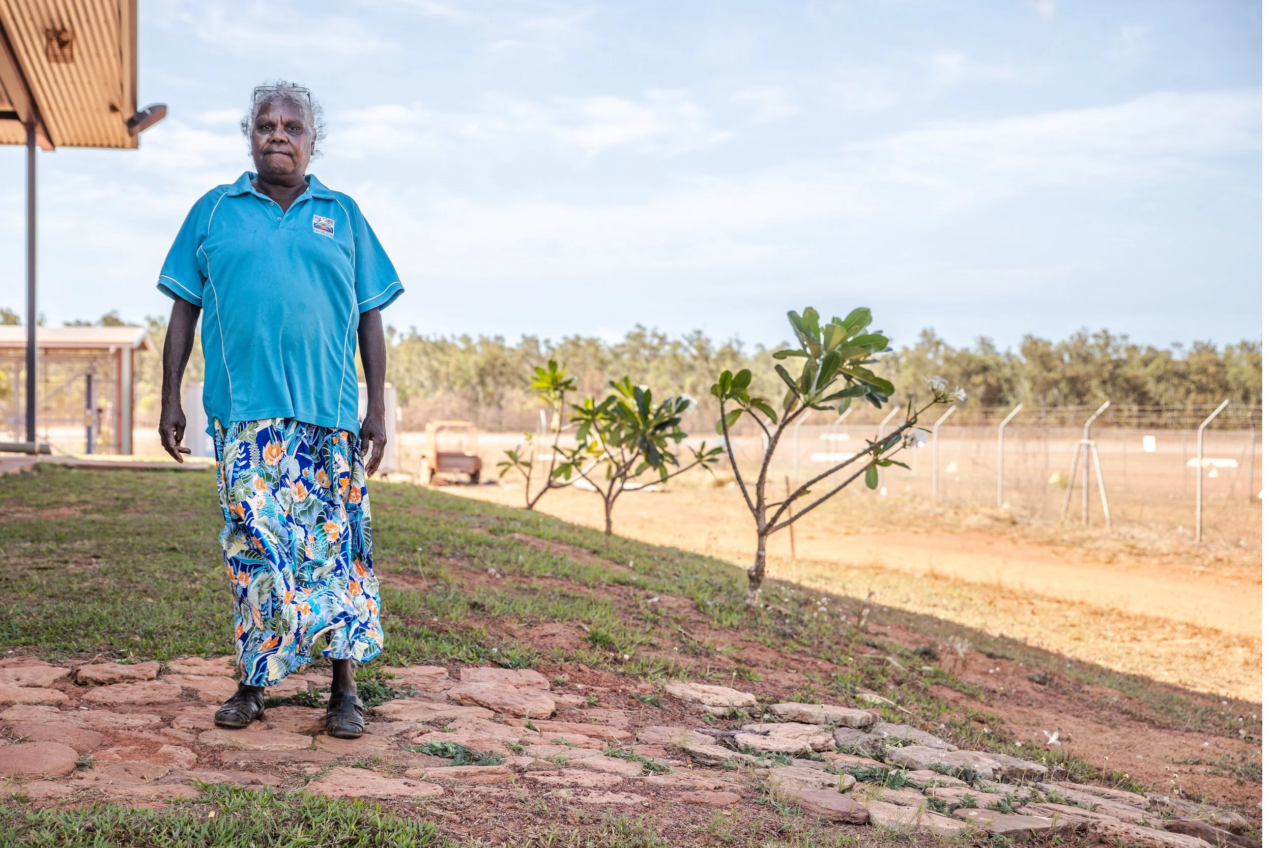 An elderly man wearing a blue polo shirt, colorful patterned wide-leg pants, and black shoes walking along a cobblestone path outdoors. There's a small tree with green leaves and white flowers, a fenced area in the background, and a clear sky with some clouds.
