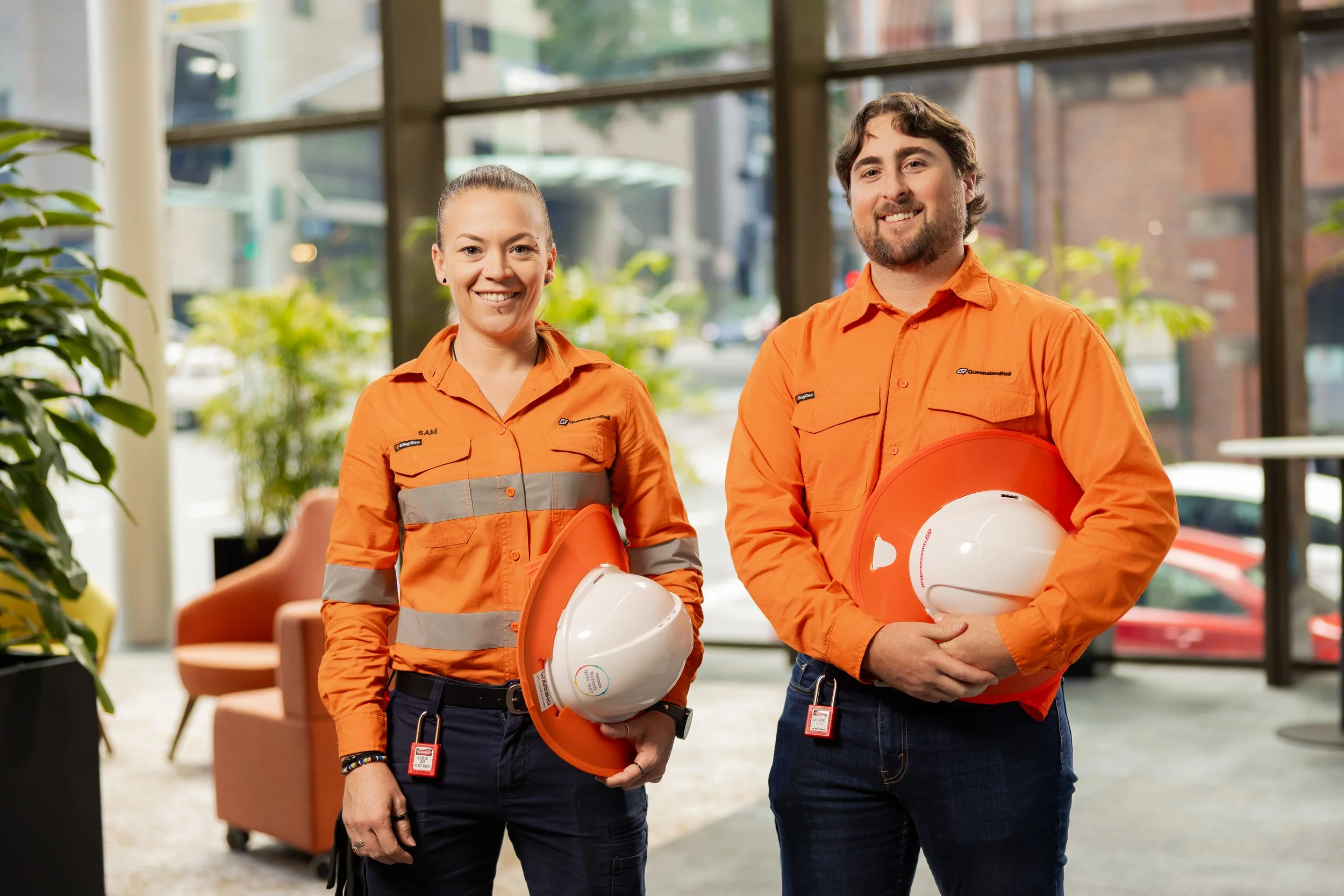 Two construction workers in orange shirts standing indoors with safety helmets, smiling at the camera.