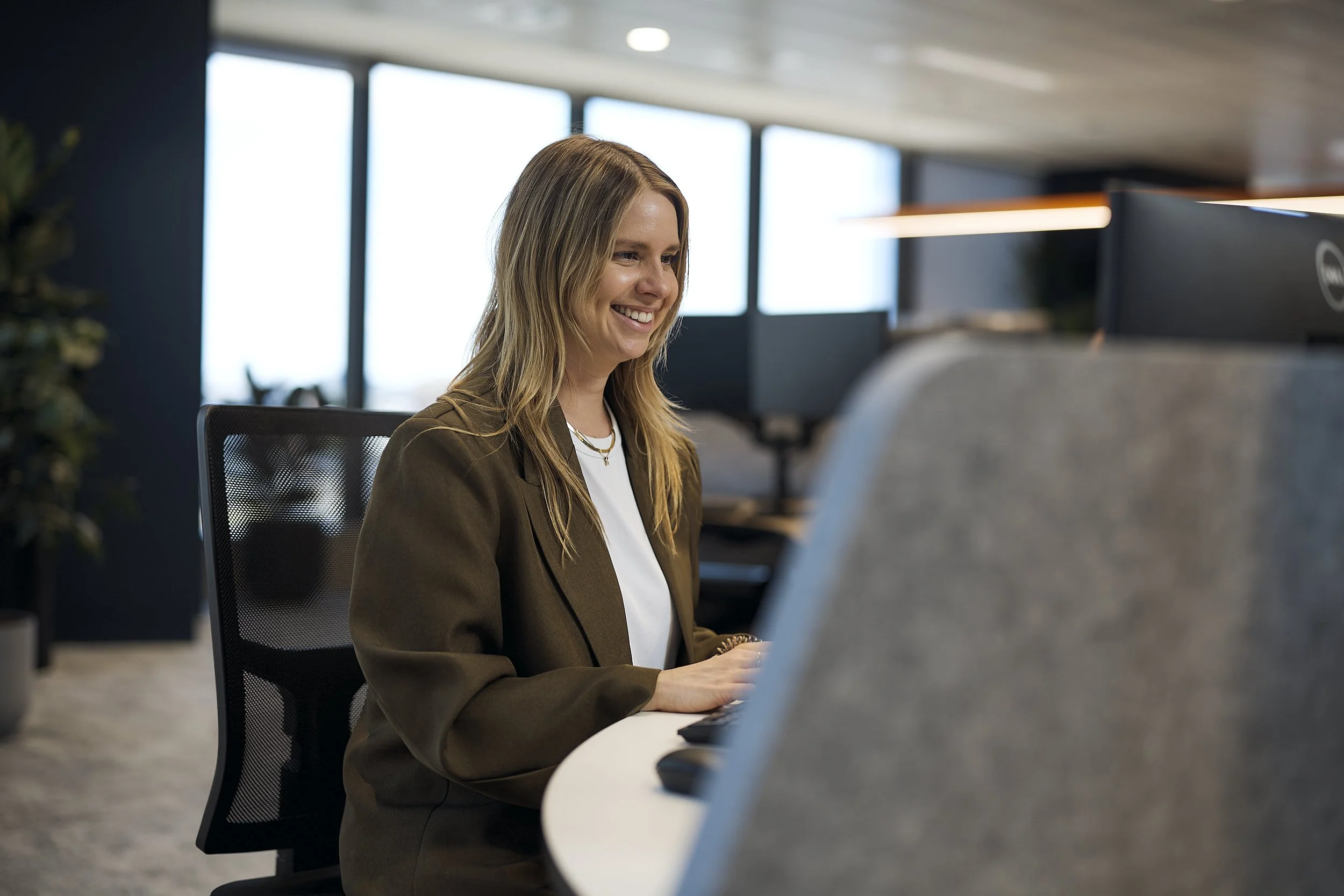 A woman with long blonde hair smiling while working at a desk in a modern office setting, with large windows in the background.
