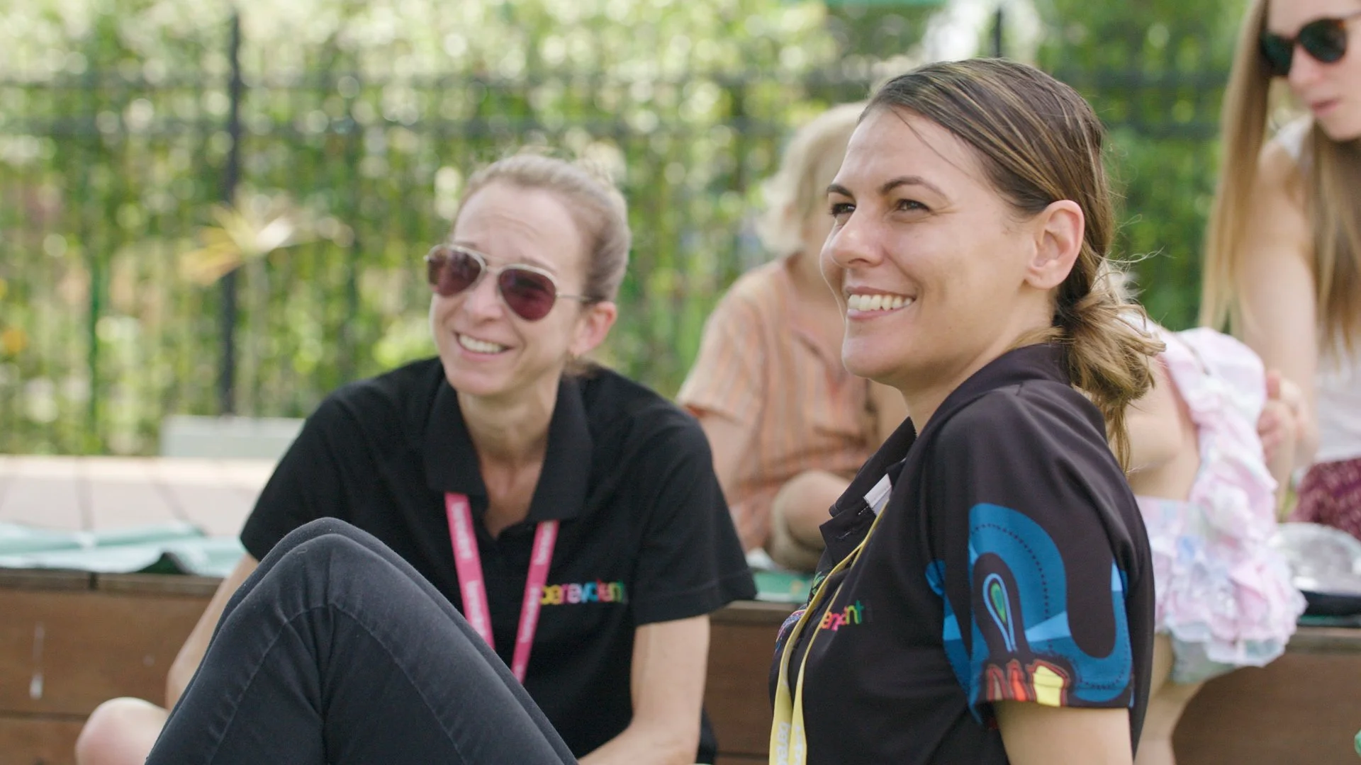 Two women sitting outdoors, smiling and chatting during a sunny day with green trees in the background. The woman on the left wears sunglasses and a black shirt, and the woman on the right has brown hair and is dressed in a colorful sports jersey.
