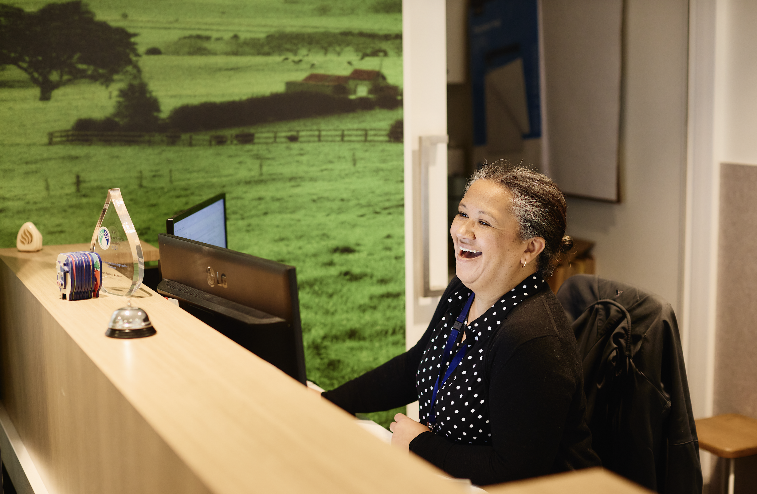 A woman with curly hair and earrings smiling while sitting at a reception desk, with a large green farm scene wall art behind her.