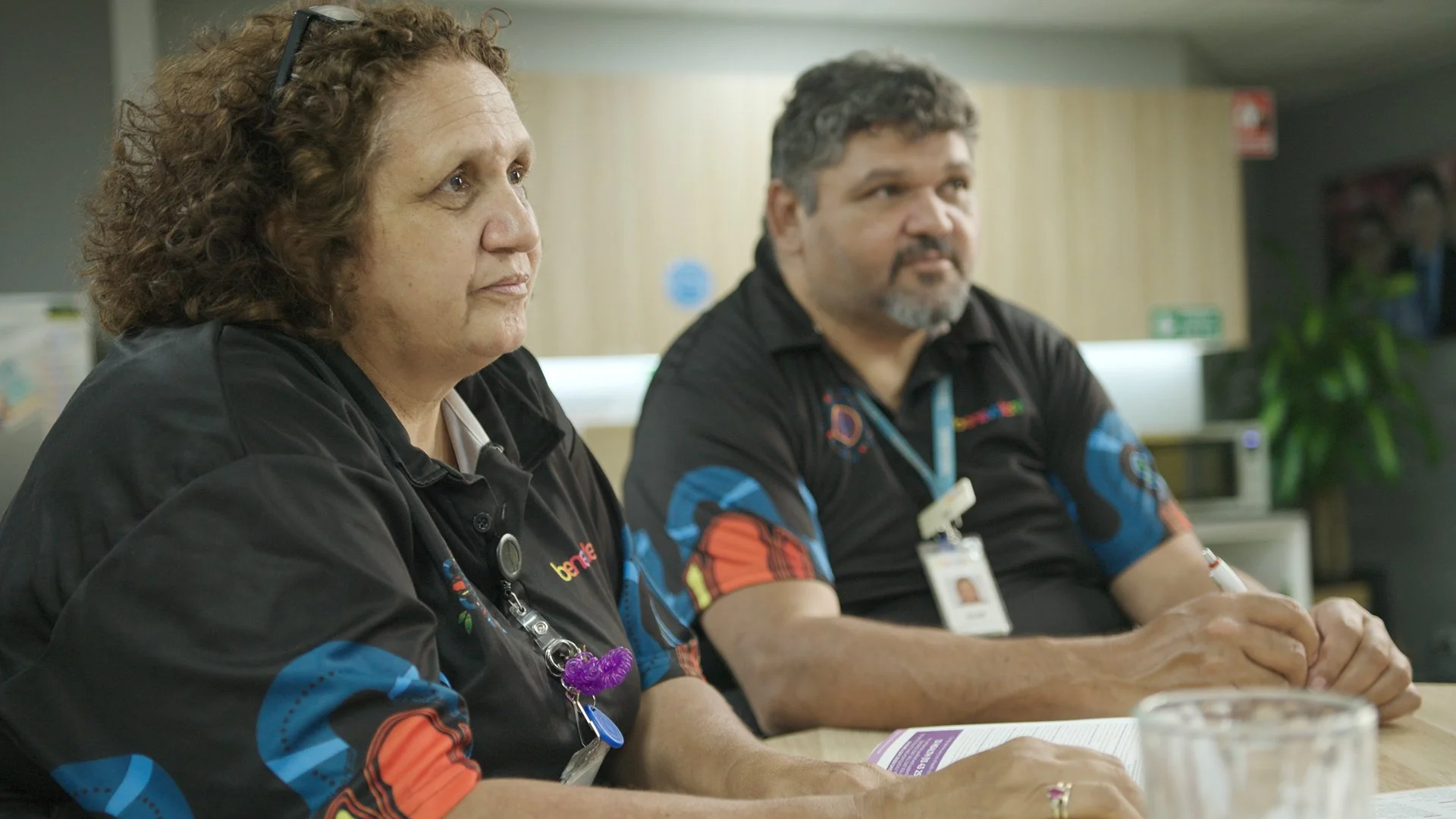 Two healthcare workers wearing black shirts with colorful patterns, sitting at a table, focused, with papers and pens in front of them.