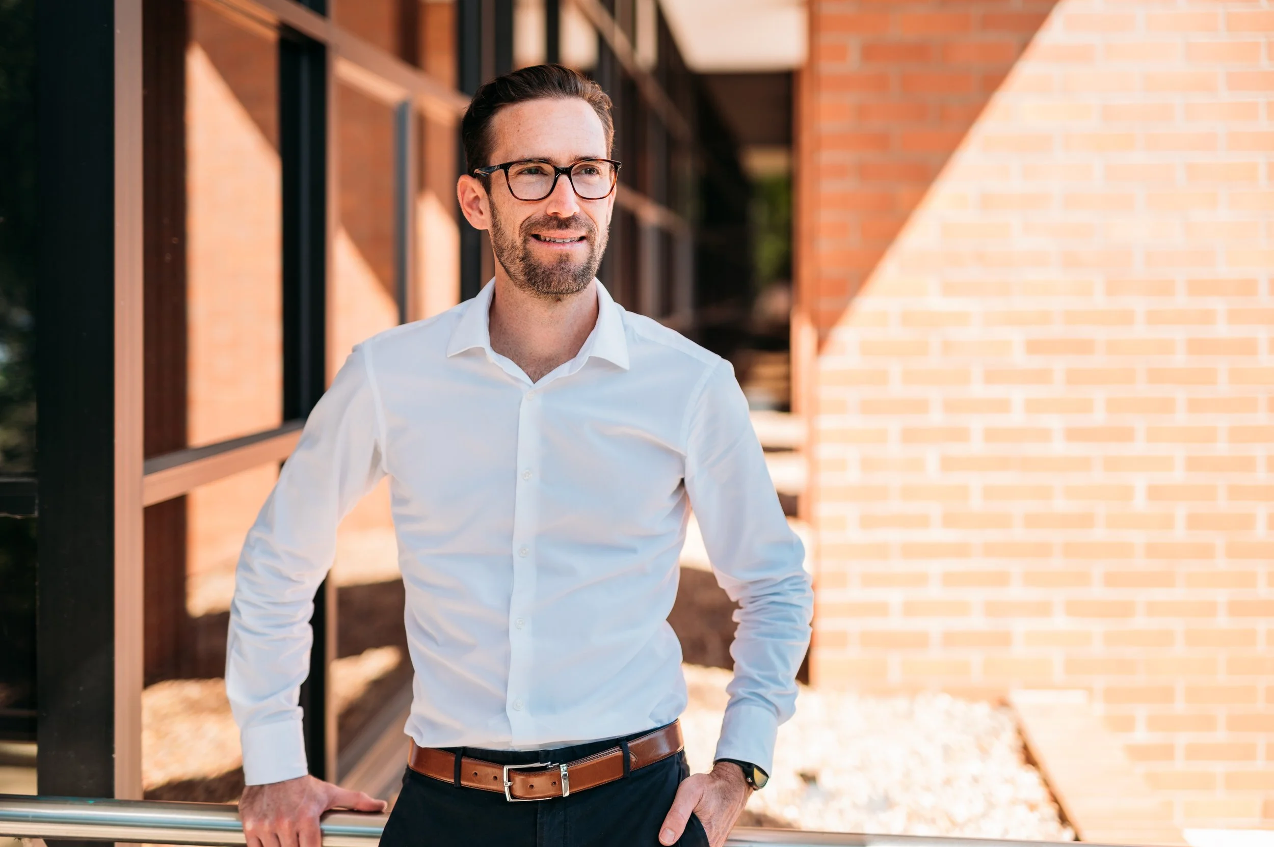 A man with dark hair, glasses, and a beard wearing a white shirt, standing outdoors near a brick building, with his right hand resting on a railing.