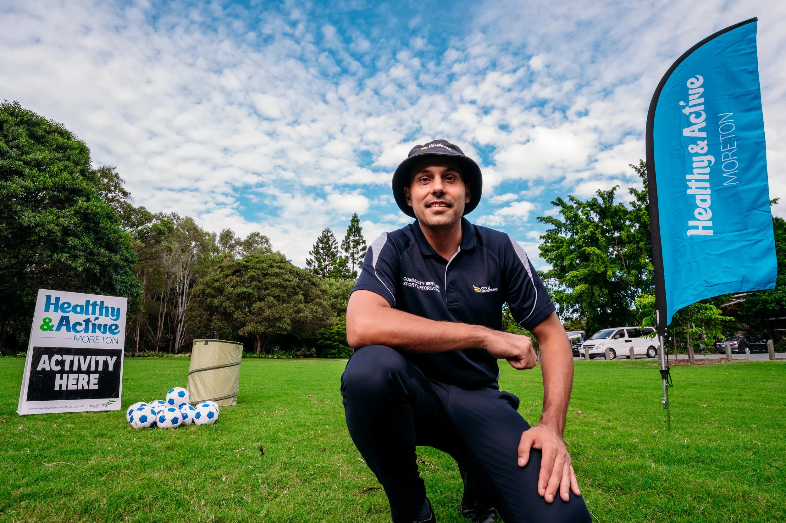 Man kneeling outdoors at a community health event, wearing a black hat and navy polo shirt, with a sign that reads 'Healthy & Active Moreton' and a flag that says 'Health & Active Moreton' in the background, with trees and a partly cloudy sky.