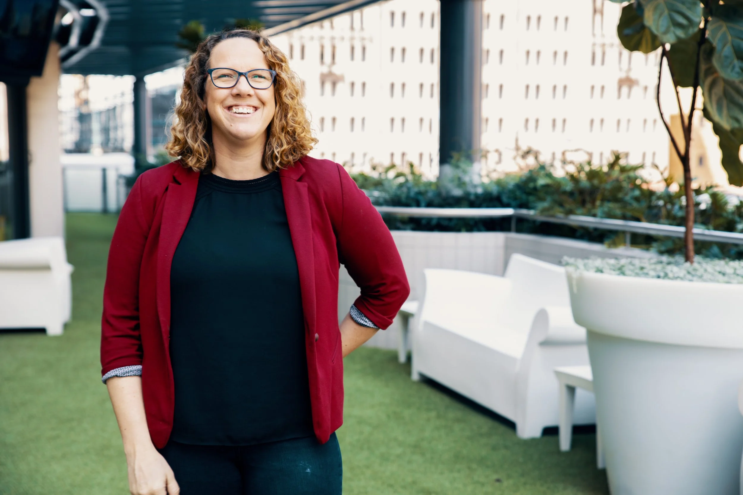 A smiling woman with curly hair and glasses wearing a red blazer and black top standing outdoors near white chairs and large potted plants in a modern urban setting.