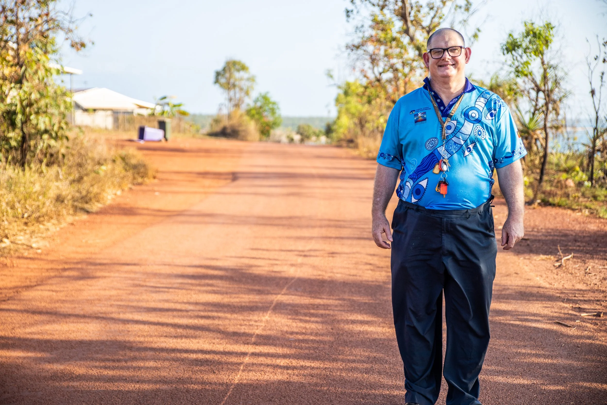 A man wearing glasses and a blue patterned shirt walking outdoors on a dirt path with trees and a house in the background.