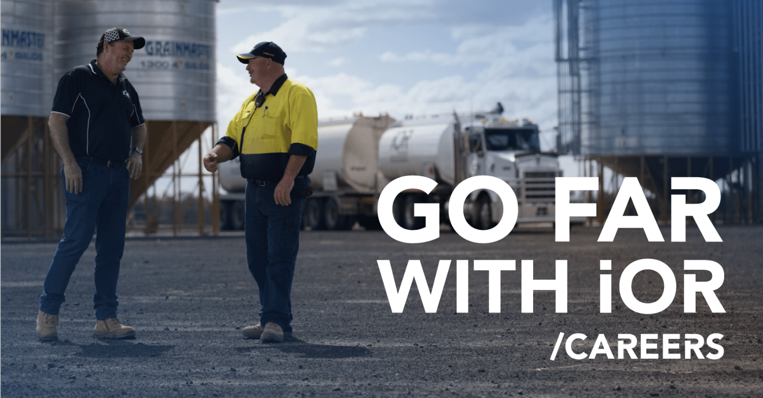 Two men in work uniforms talking outdoors near storage tanks and a semi-truck on a dirt lot on a partly cloudy day.