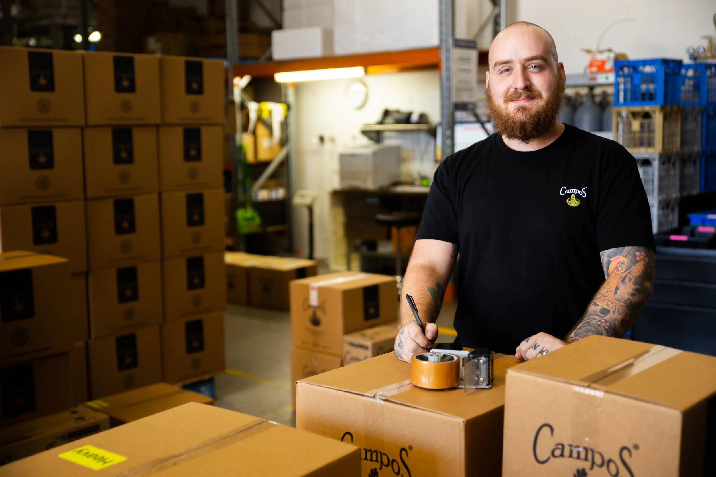 A man with a beard and tattoos on his arms wearing a black T-shirt with a logo is standing in a warehouse surrounded by stacked boxes. He is holding a marker and is about to tape a box.