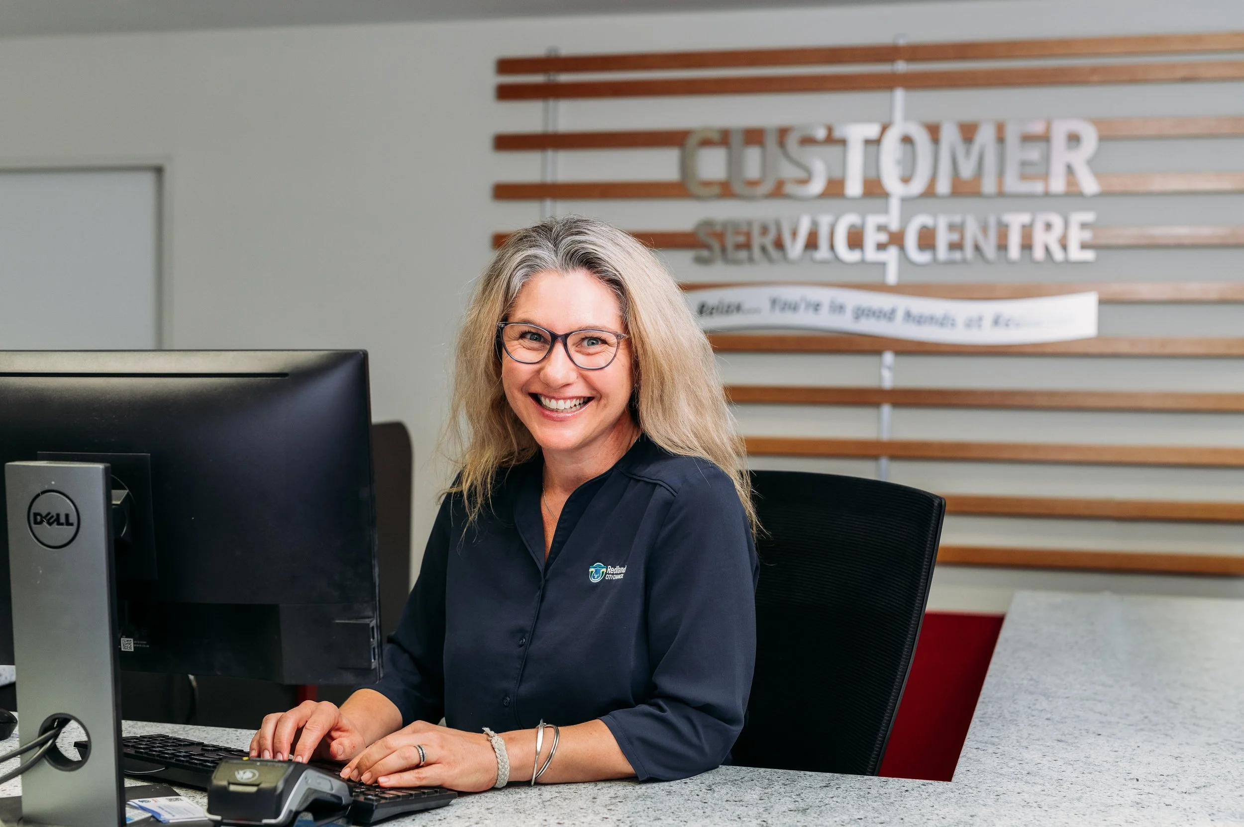 A smiling woman with long blond hair and glasses sitting at a desk in a customer service center. Behind her is a sign that reads "Customer Service Centre" with wooden slats on the wall.