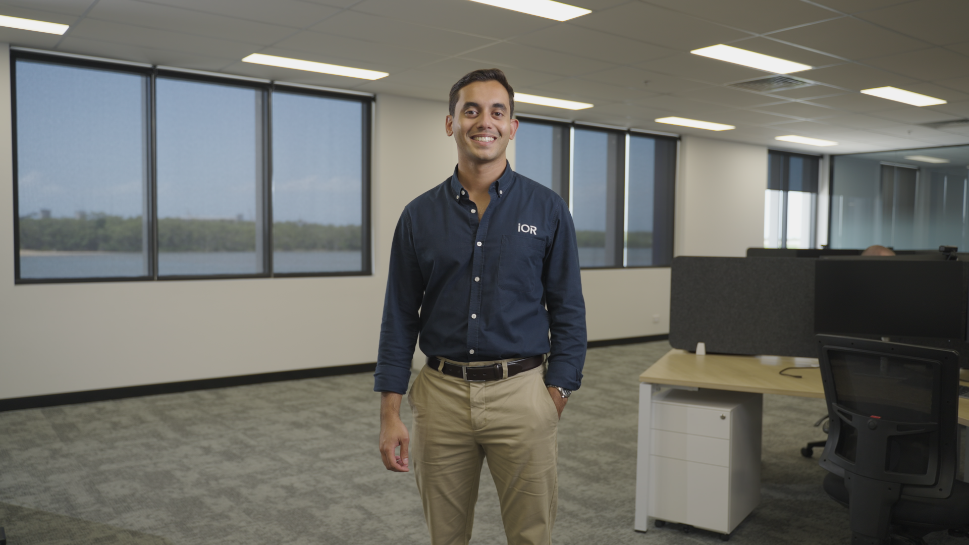 A young man smiling and standing in an office with large windows overlooking a body of water.