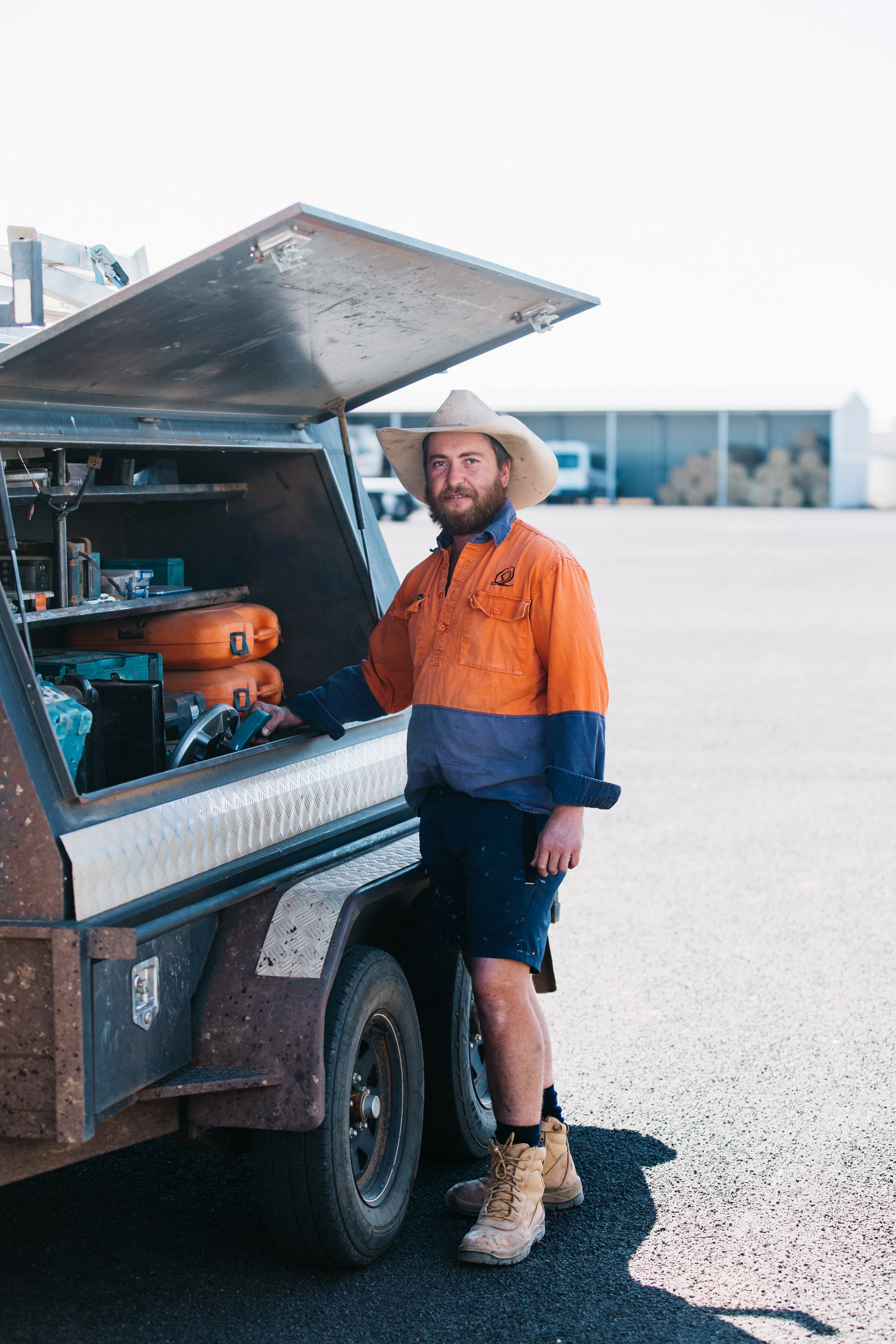 A man wearing a wide-brimmed hat, orange work shirt, and beige work boots stands next to a utility vehicle with its storage compartment open, in an outdoor setting with a large industrial or storage area in the background.