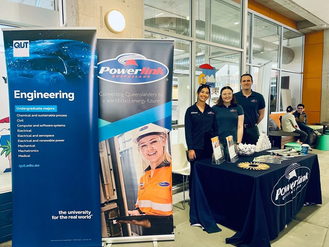 Three people standing behind a table at a promotional booth with banners for Queensland University of Technology and Powerlink Queensland, inside a modern building with large windows and other people sitting and talking in the background.