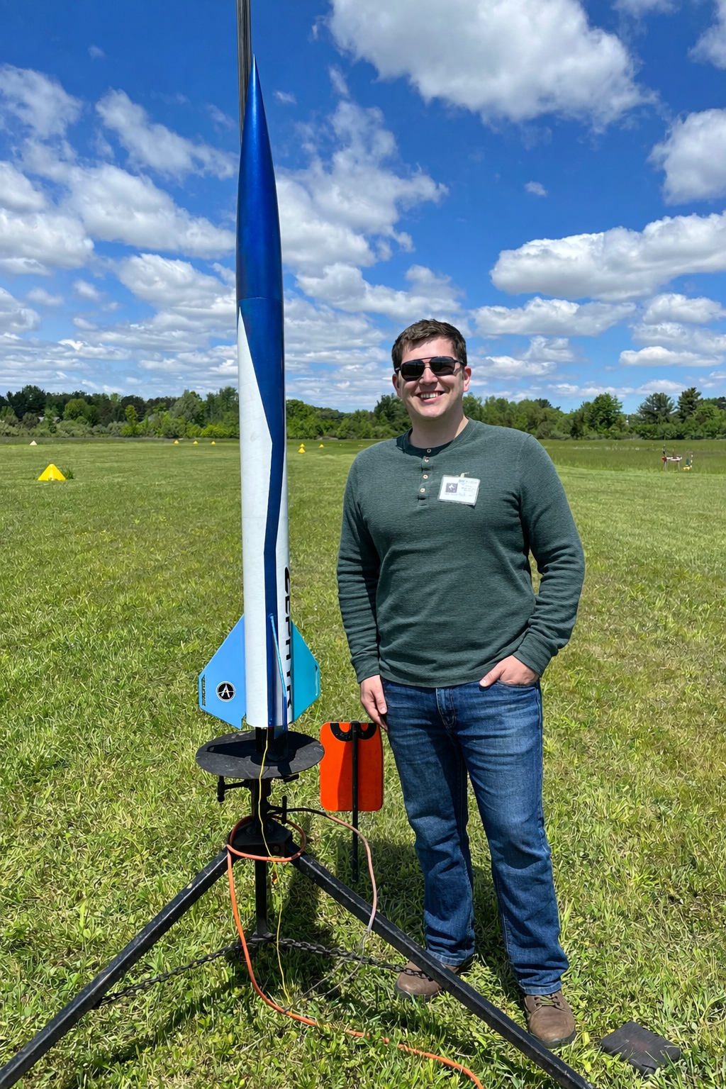 A smiling man wearing sunglasses and a gray shirt standing outdoors on a grassy field next to a large model rocket on a launch pad, with a partly cloudy sky and trees in the background.