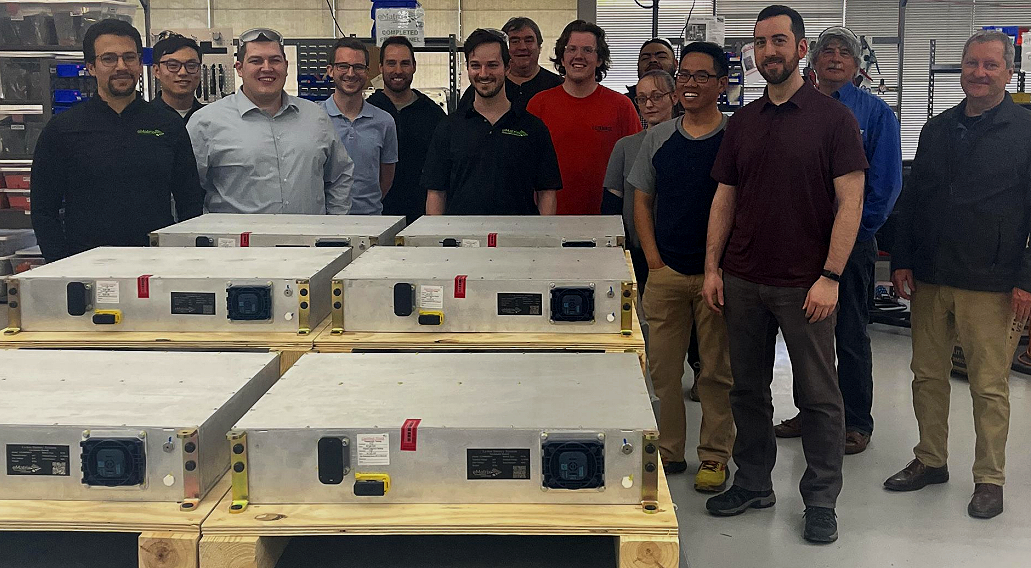 Group of people standing in a workshop behind several large metal boxes on wooden pallets.